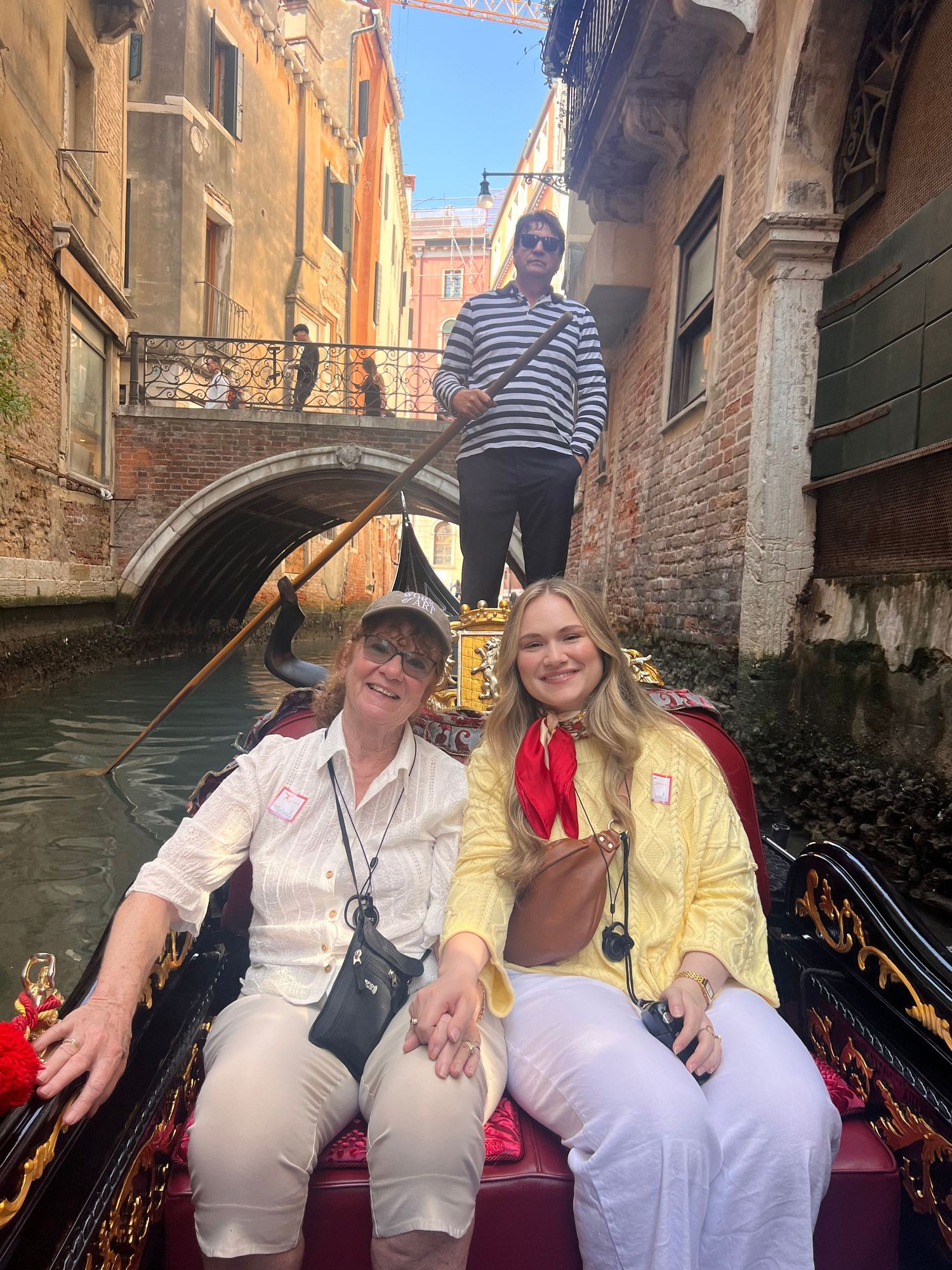 Two women smile on a gondola ride through a narrow Venice canal with a gondolier standing behind them.