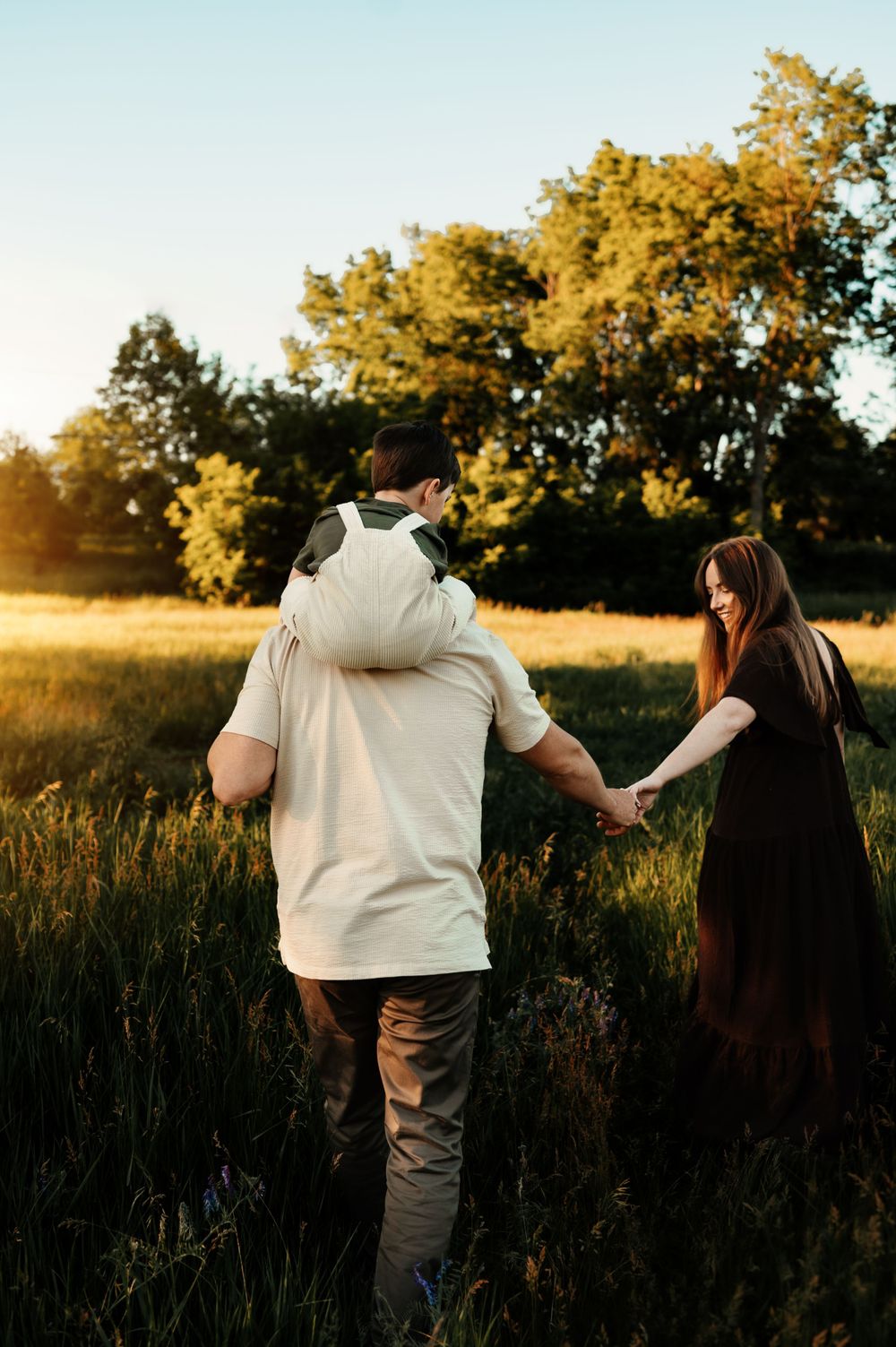 A couple holds hands while walking through a sun-lit meadow with golden trees in the background.