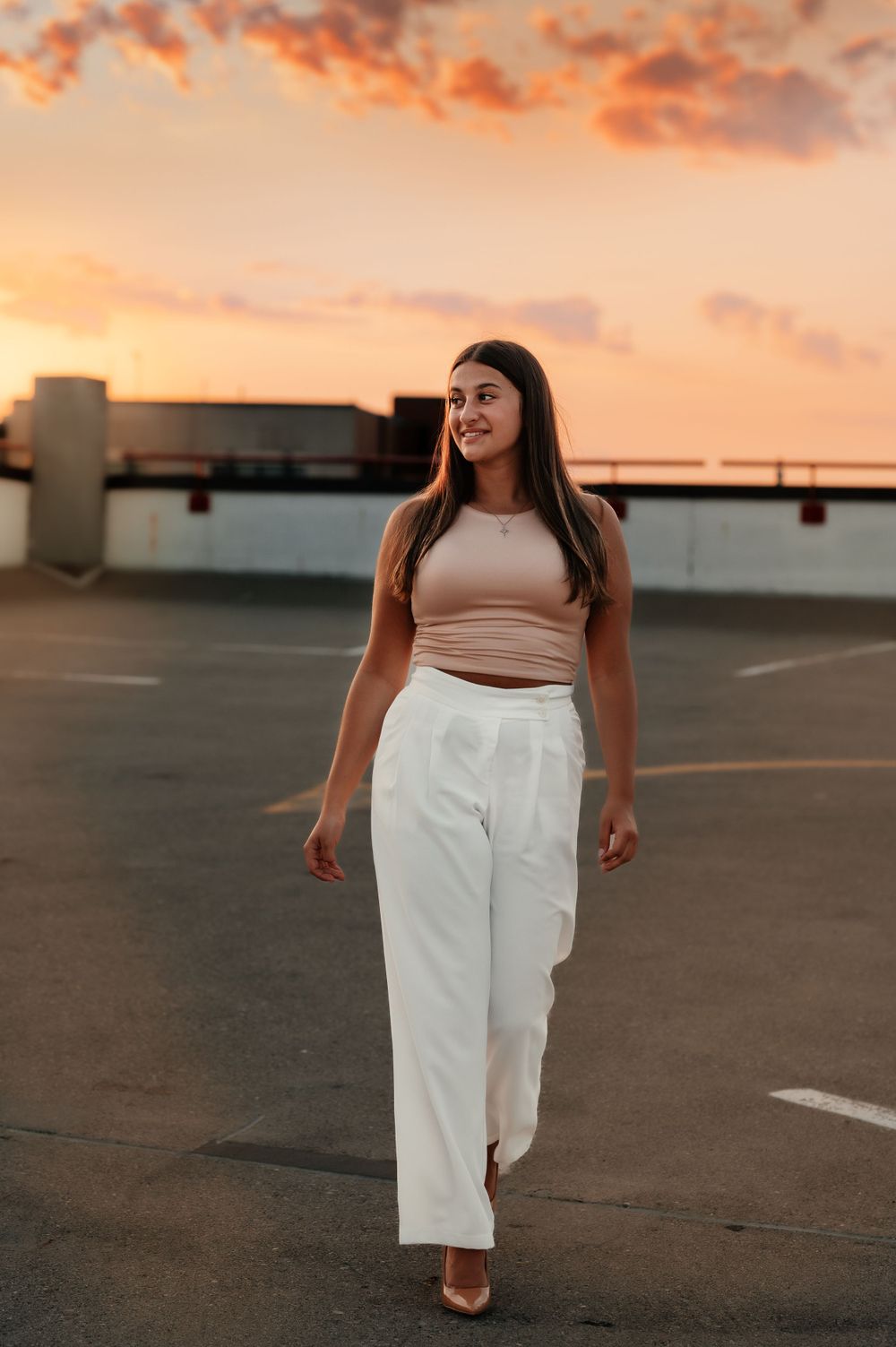 A woman in a crop top and white pants walks across a rooftop parking lot during a colorful sunset.