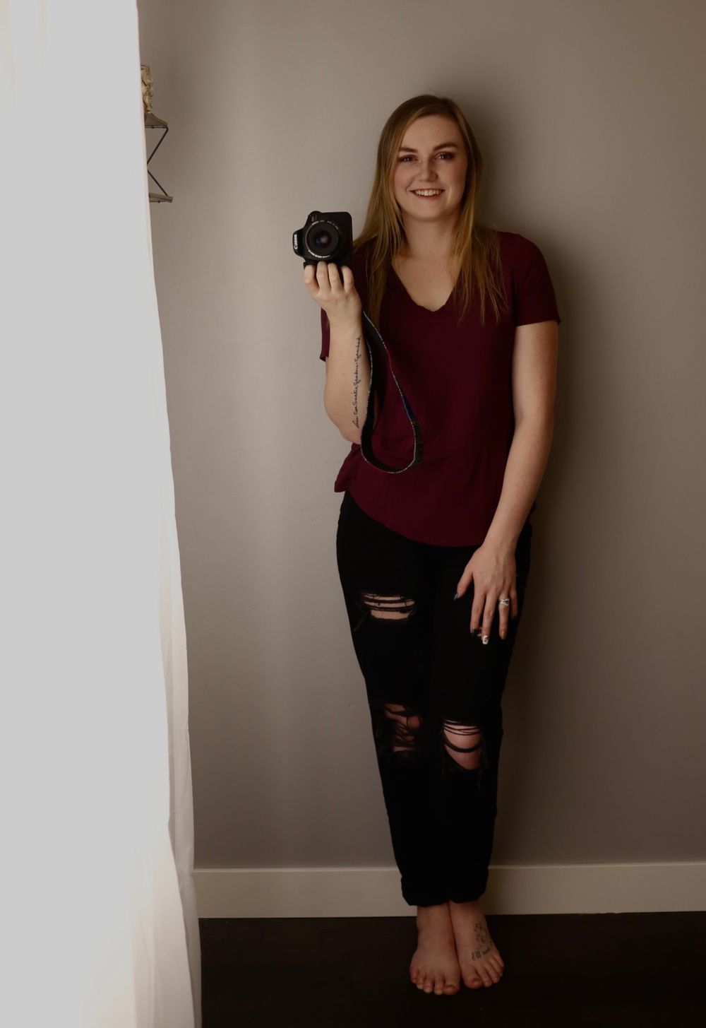 Photographer in burgundy shirt and ripped jeans holds a camera while standing barefoot against a neutral wall.