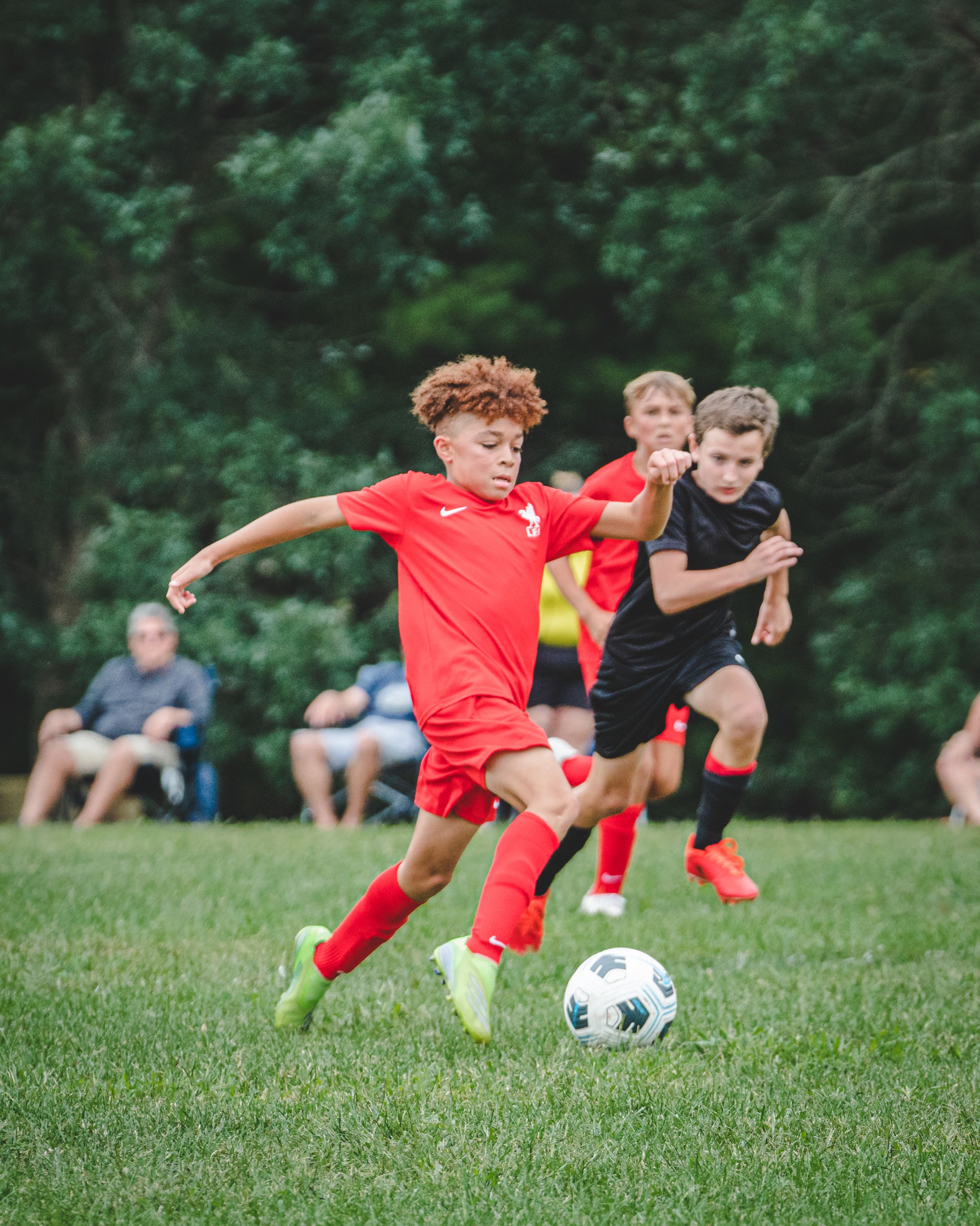 Young boy in red uniform dribbles soccer ball during outdoor youth league match.
