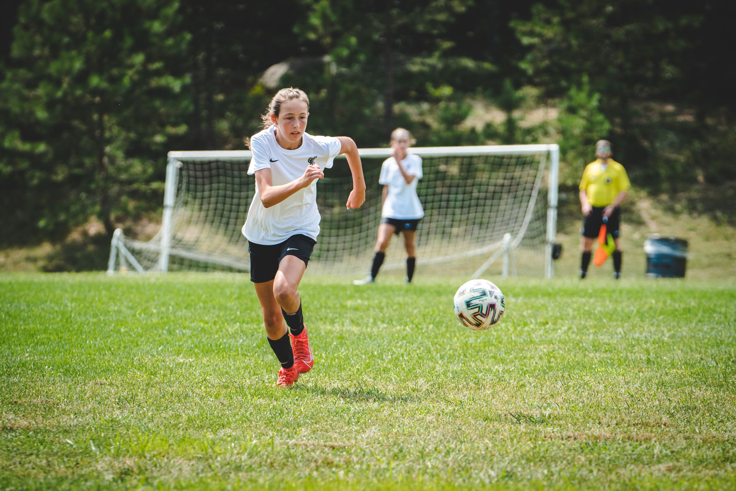 Young girl in white uniform chases soccer ball across green field with goal visible in background.