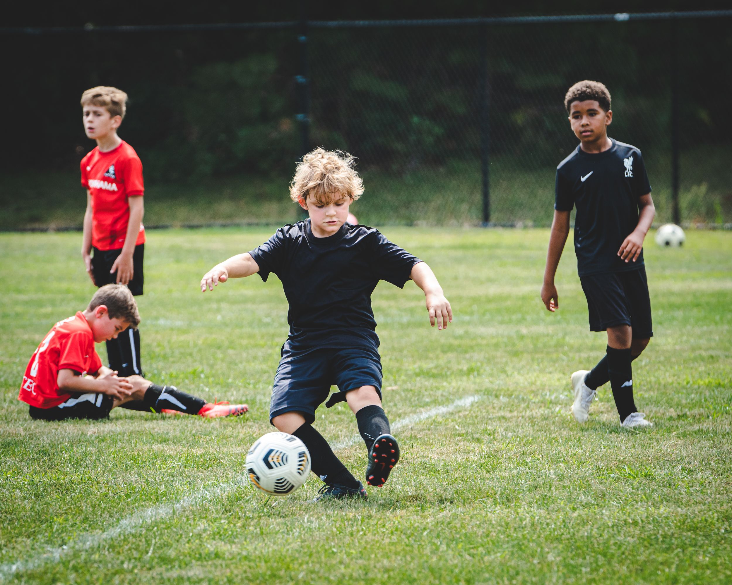 Blonde youth soccer player kicks ball during outdoor match with teammates watching nearby.
