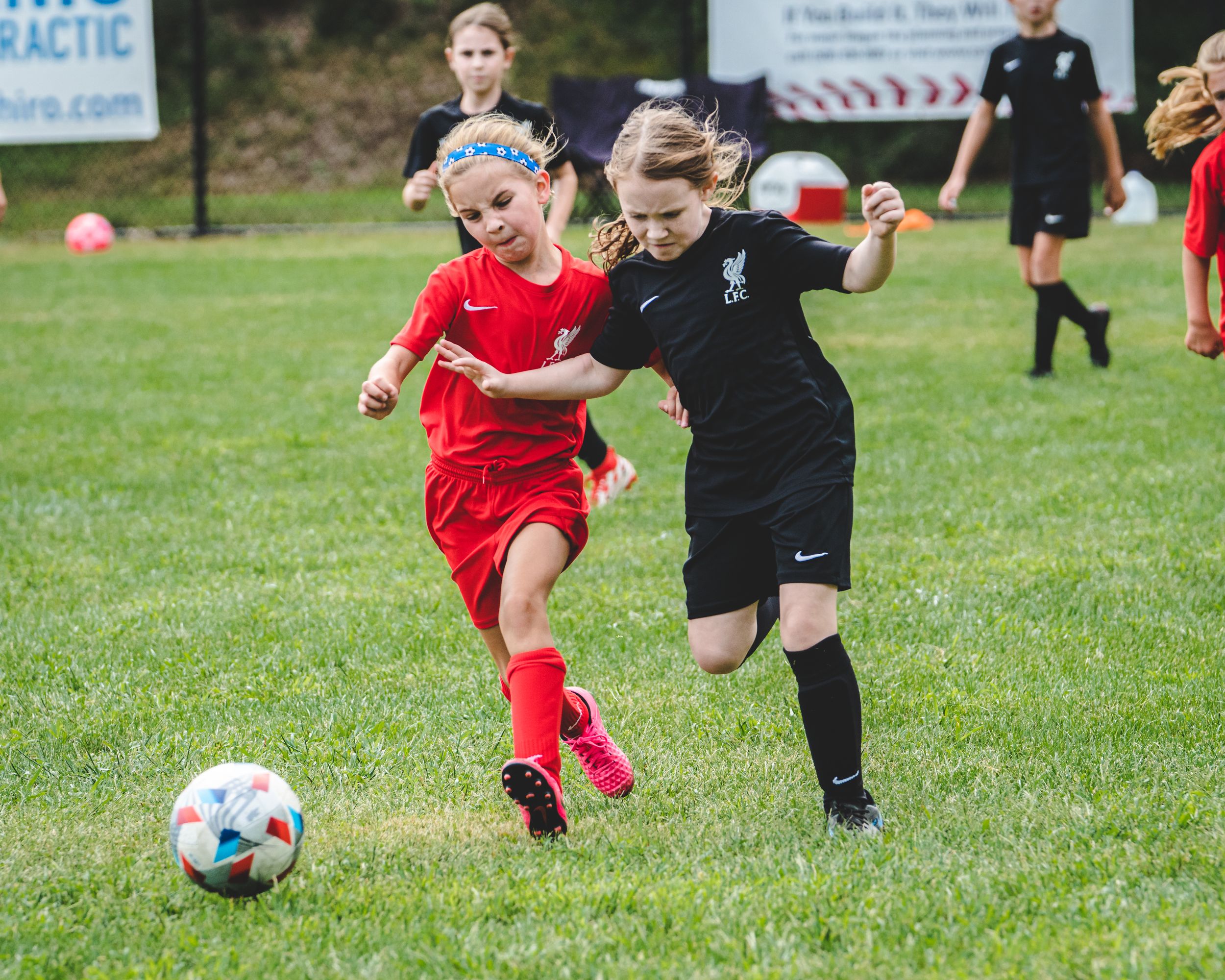 Two young girls compete for the ball during a youth soccer match, one in red and one in black uniforms.