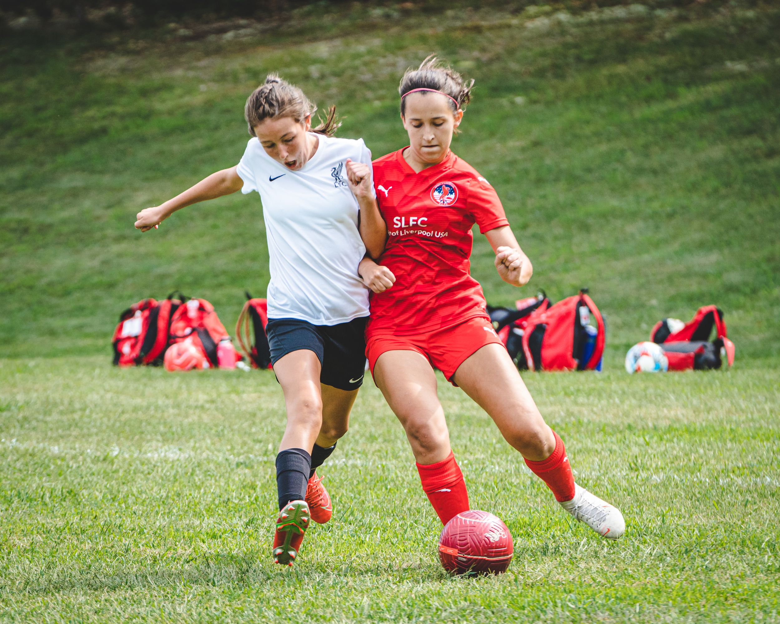Two teenage girls battle for possession during a competitive youth soccer game on a green grass field.