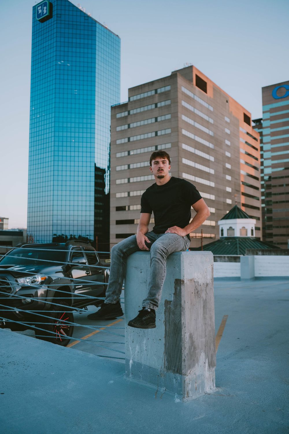 Young man in black t-shirt and gray jeans sitting on concrete pillar on urban rooftop with city skyscrapers in background.