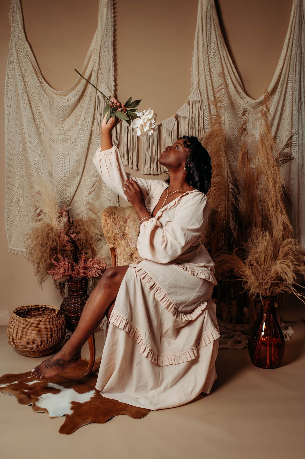 A dreamy portrait in neutral tones features a seated figure in white dress with pampas grass and macrame wall hangings.