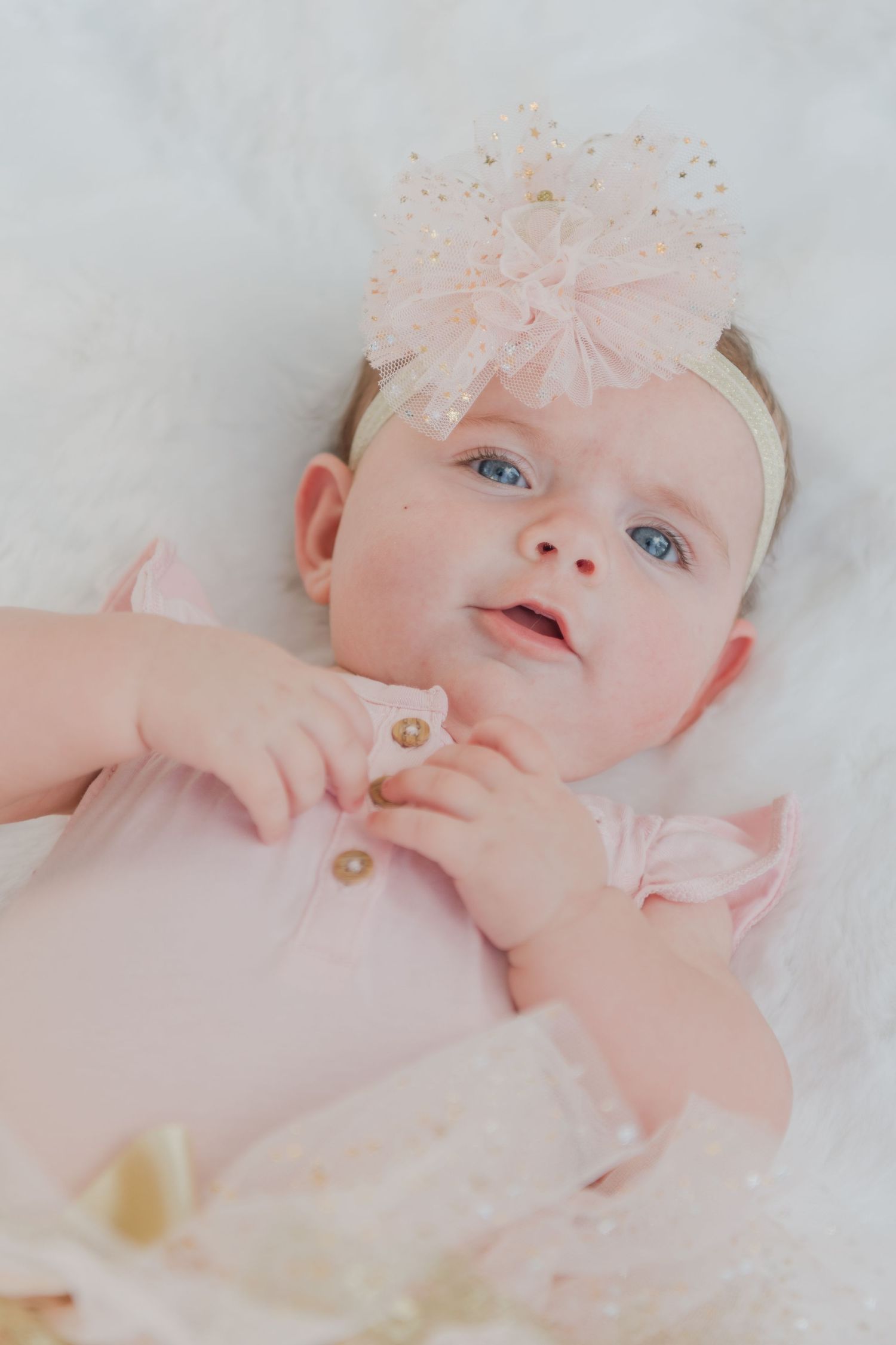 Baby wearing pink outfit and bow headband lying on white fur backdrop.