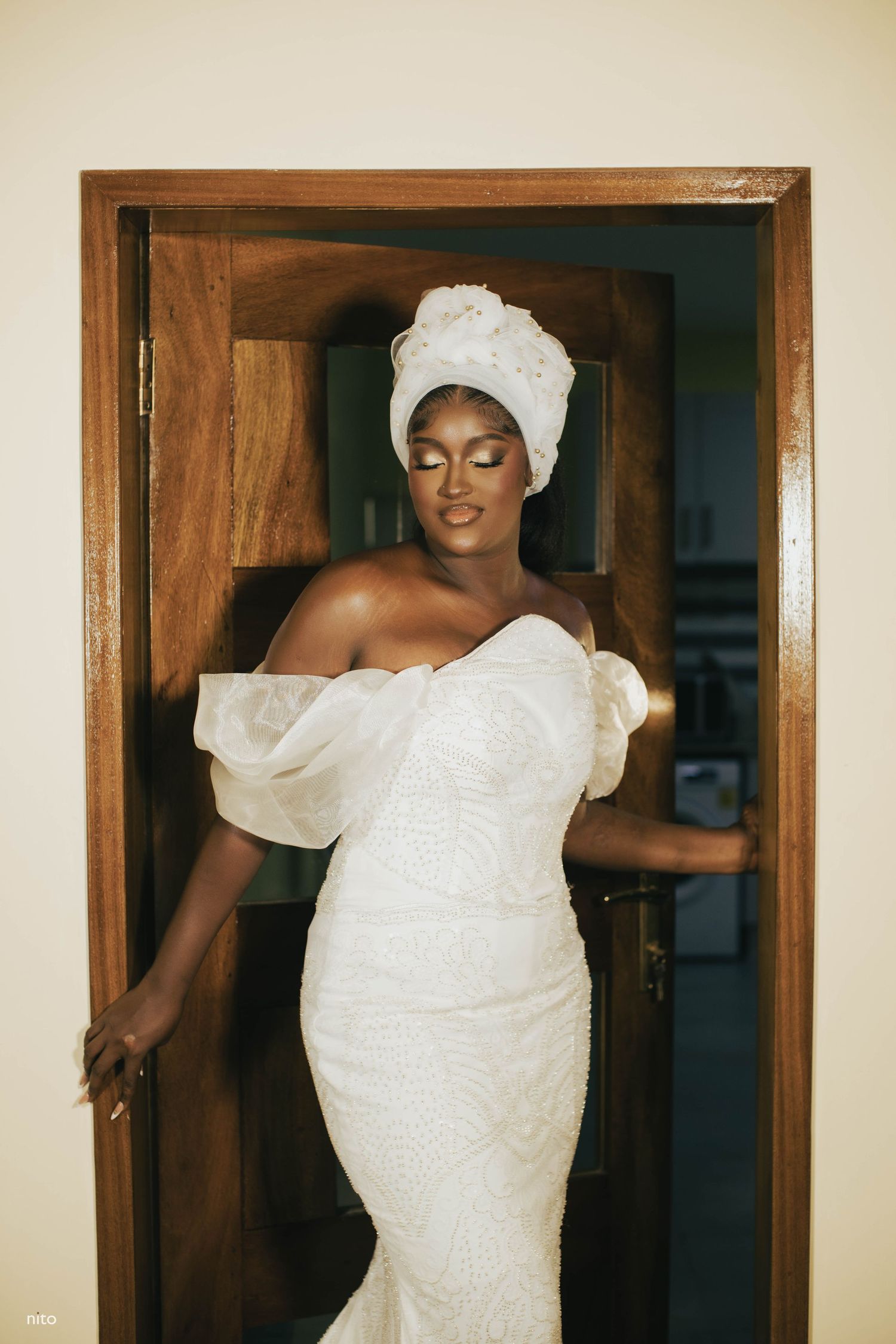 Person in off-shoulder white wedding gown and headwrap poses elegantly in doorframe.