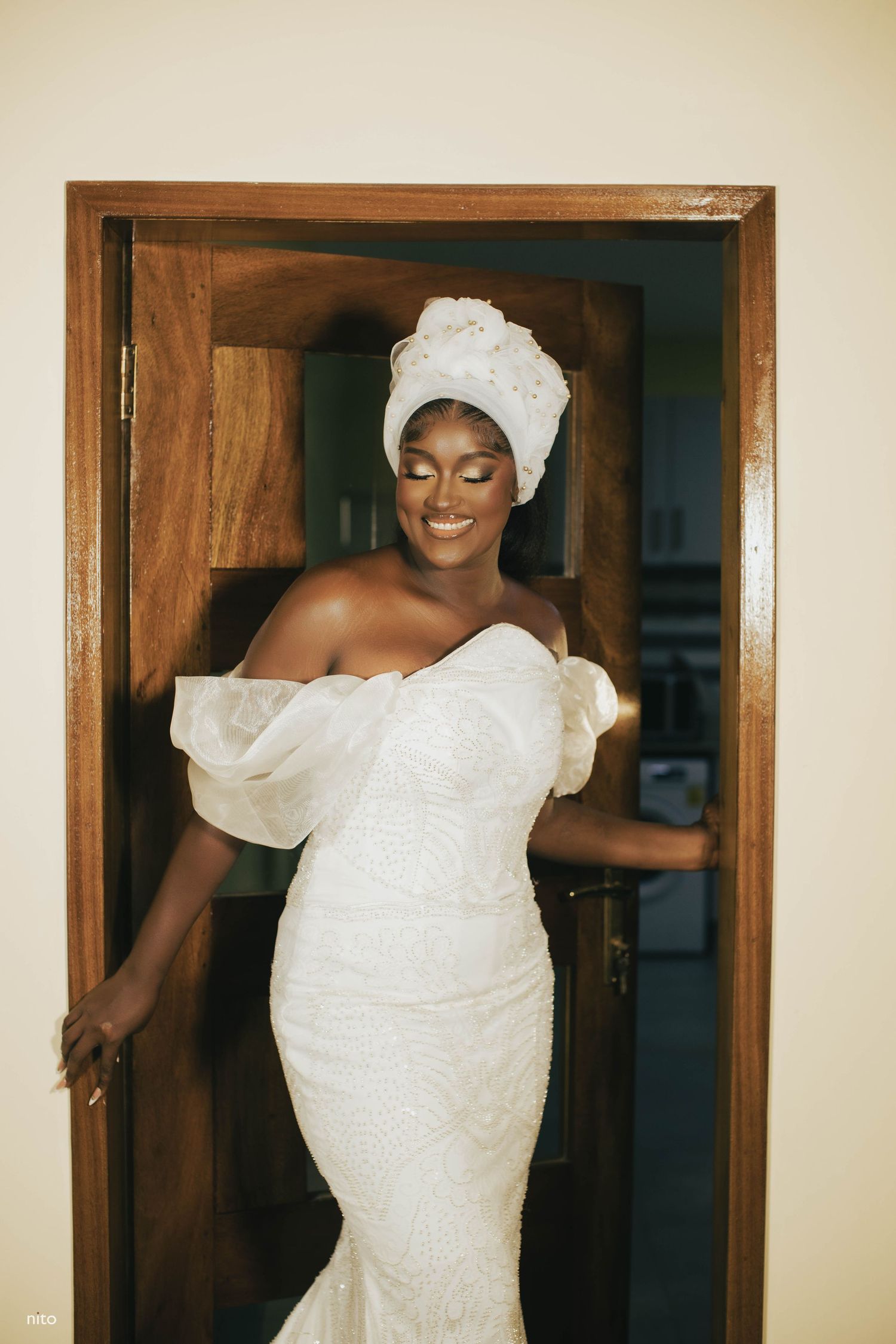 Person in white wedding dress and headwrap poses joyfully in wooden doorframe.