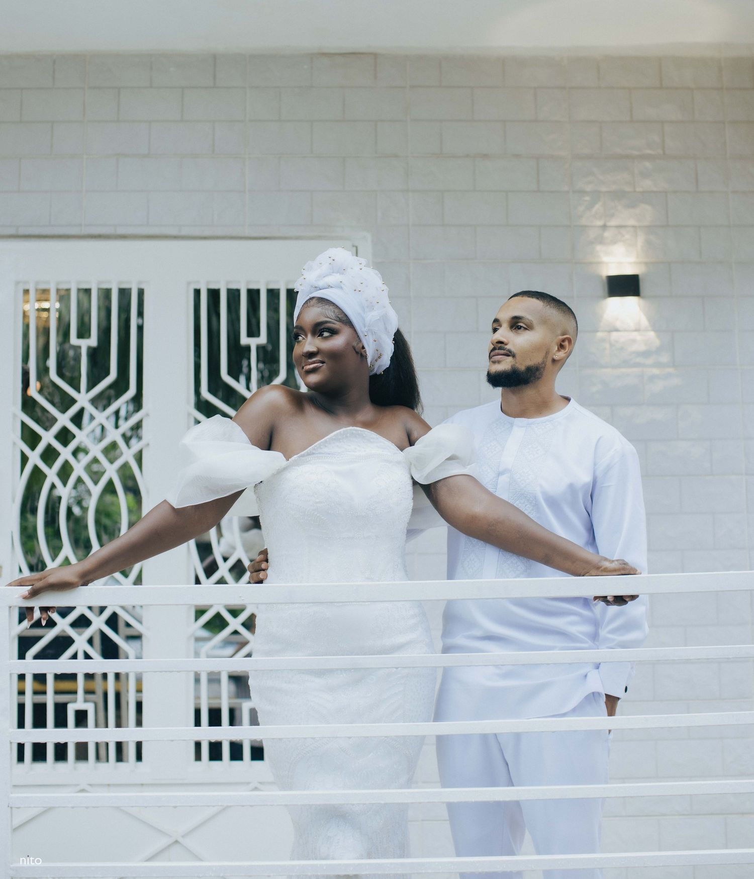 Couple in white attire pose together on balcony with decorative white railings.