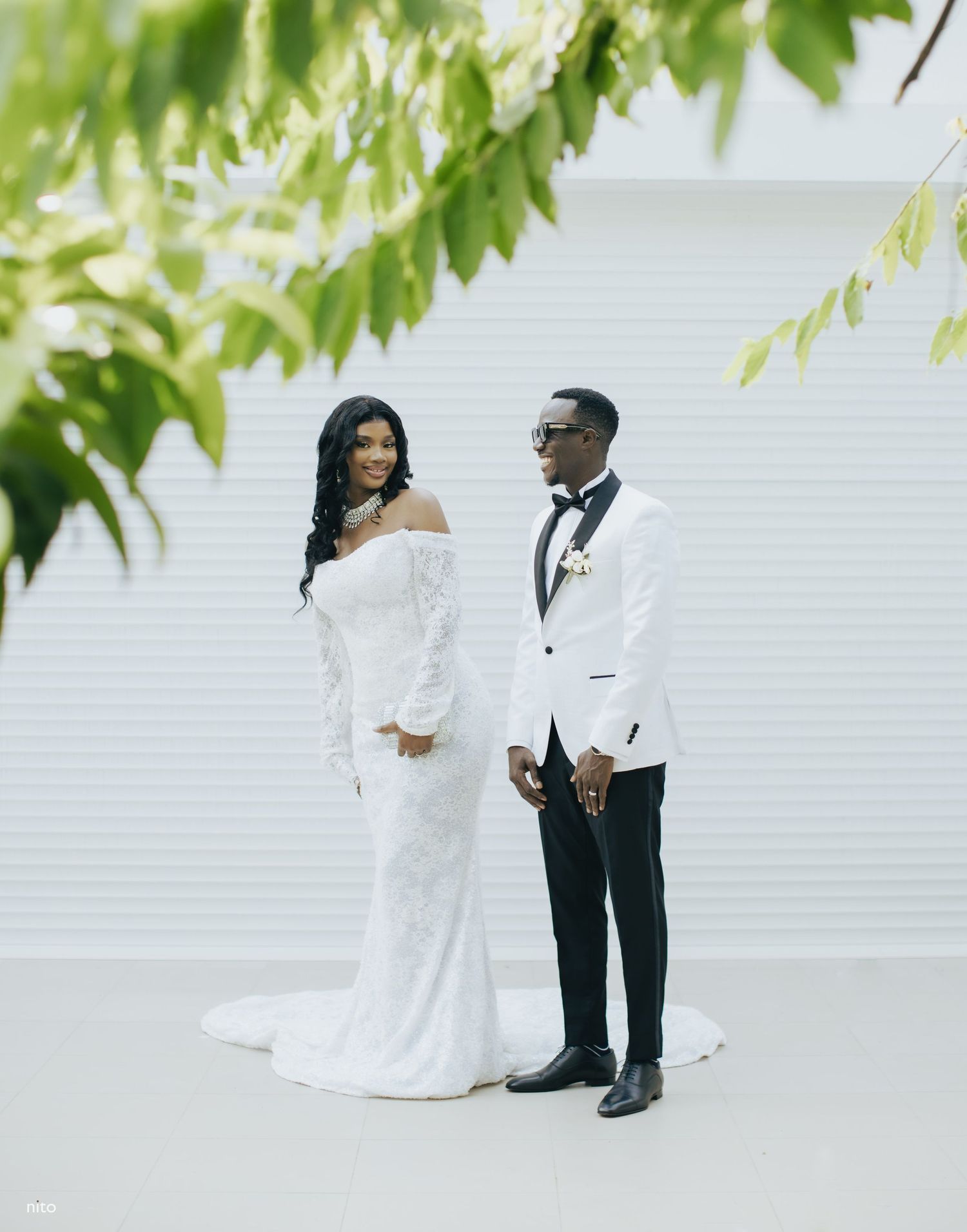 Elegant couple dressed in formal white wedding attire pose together against a white wall under green foliage.