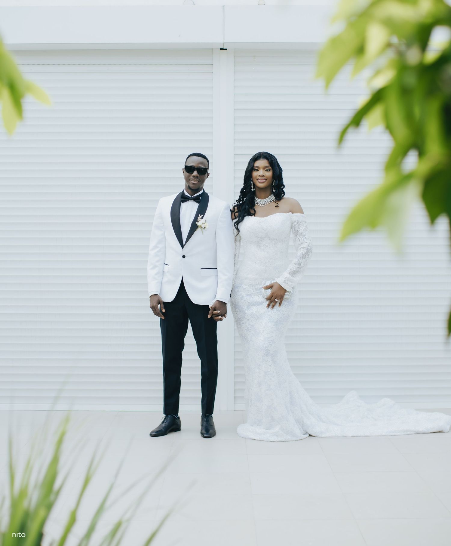 A wedding couple poses together against a white wall, wearing elegant white attire with a tropical plant framing the shot.