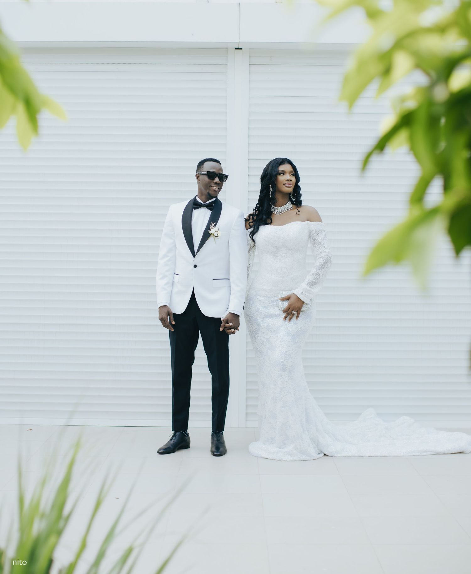 A couple poses together in formal wedding attire - white dress and white tuxedo jacket - against a white wall outdoors.