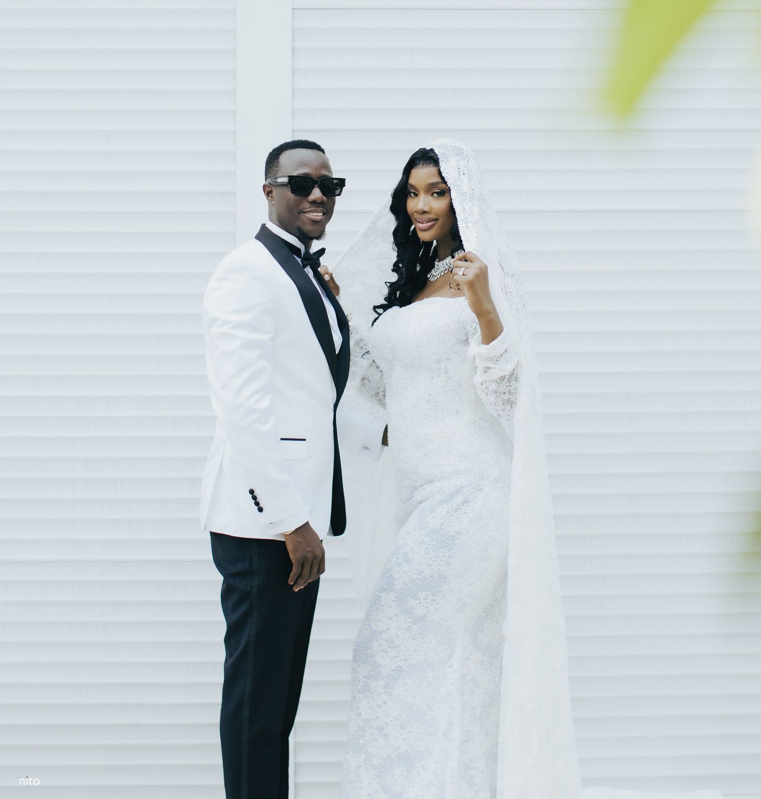 A bride and groom pose together against a white wall, with the groom in a white tuxedo and the bride in an elegant gown.