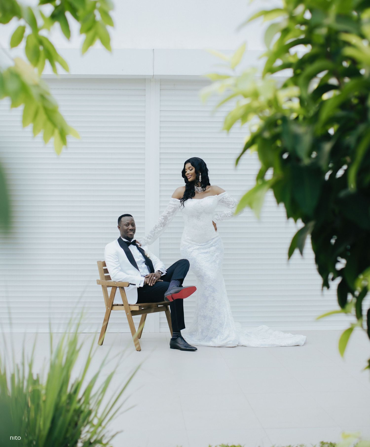 Elegant wedding photo captured through green leaves with a couple seated against a white backdrop.