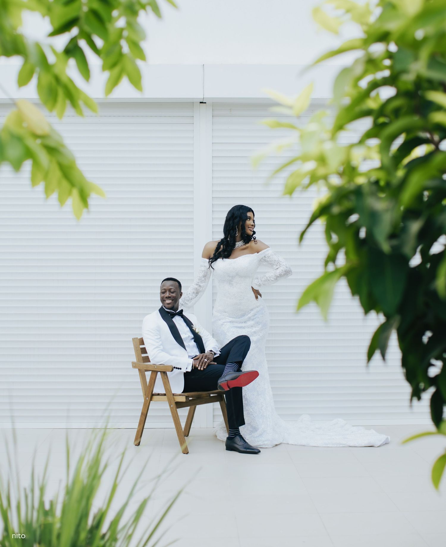 A couple poses creatively for wedding photos near greenery and a white wall with artistic styling.