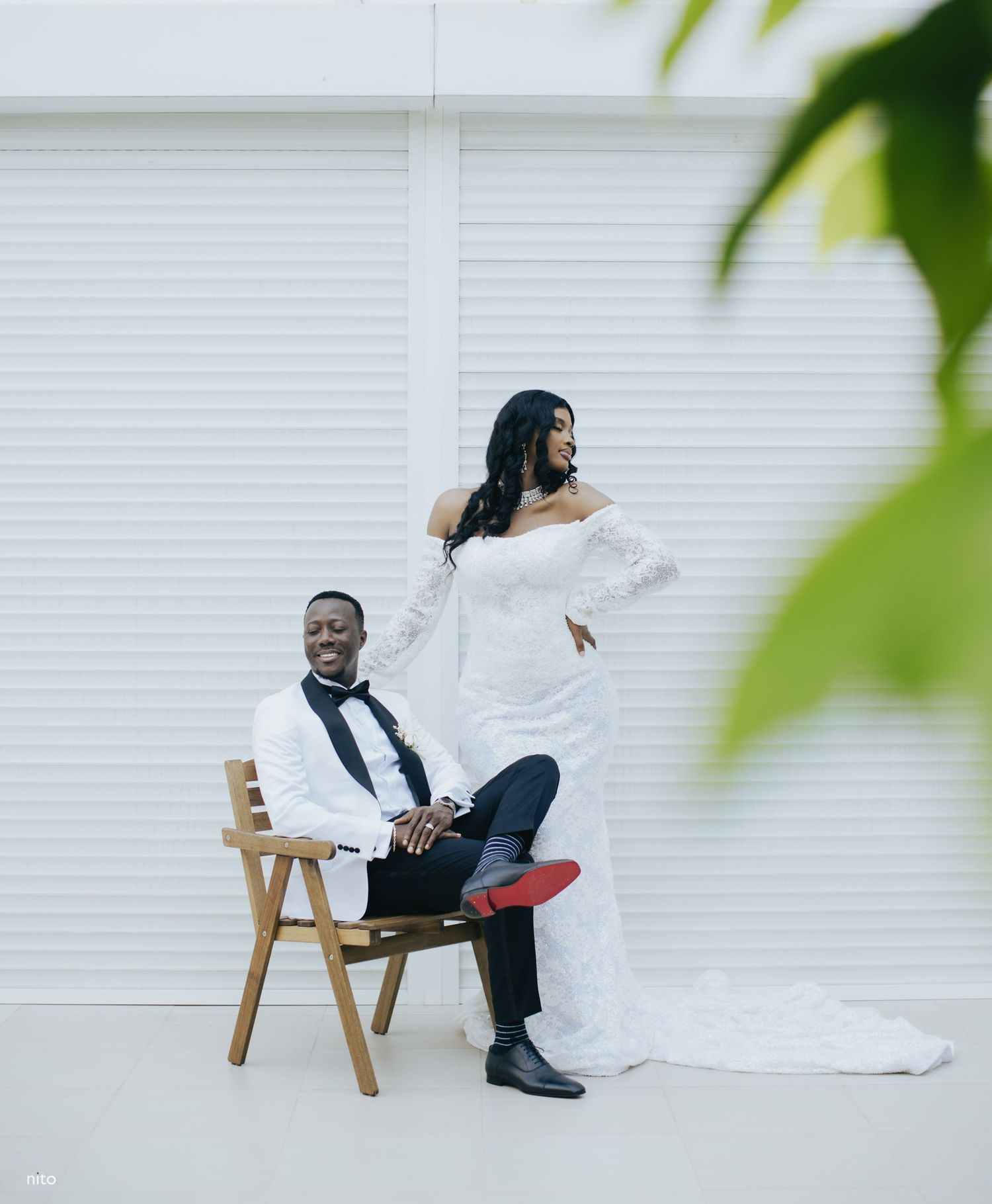 A romantic wedding photo featuring a couple in elegant white attire, with one person seated on a wooden chair.