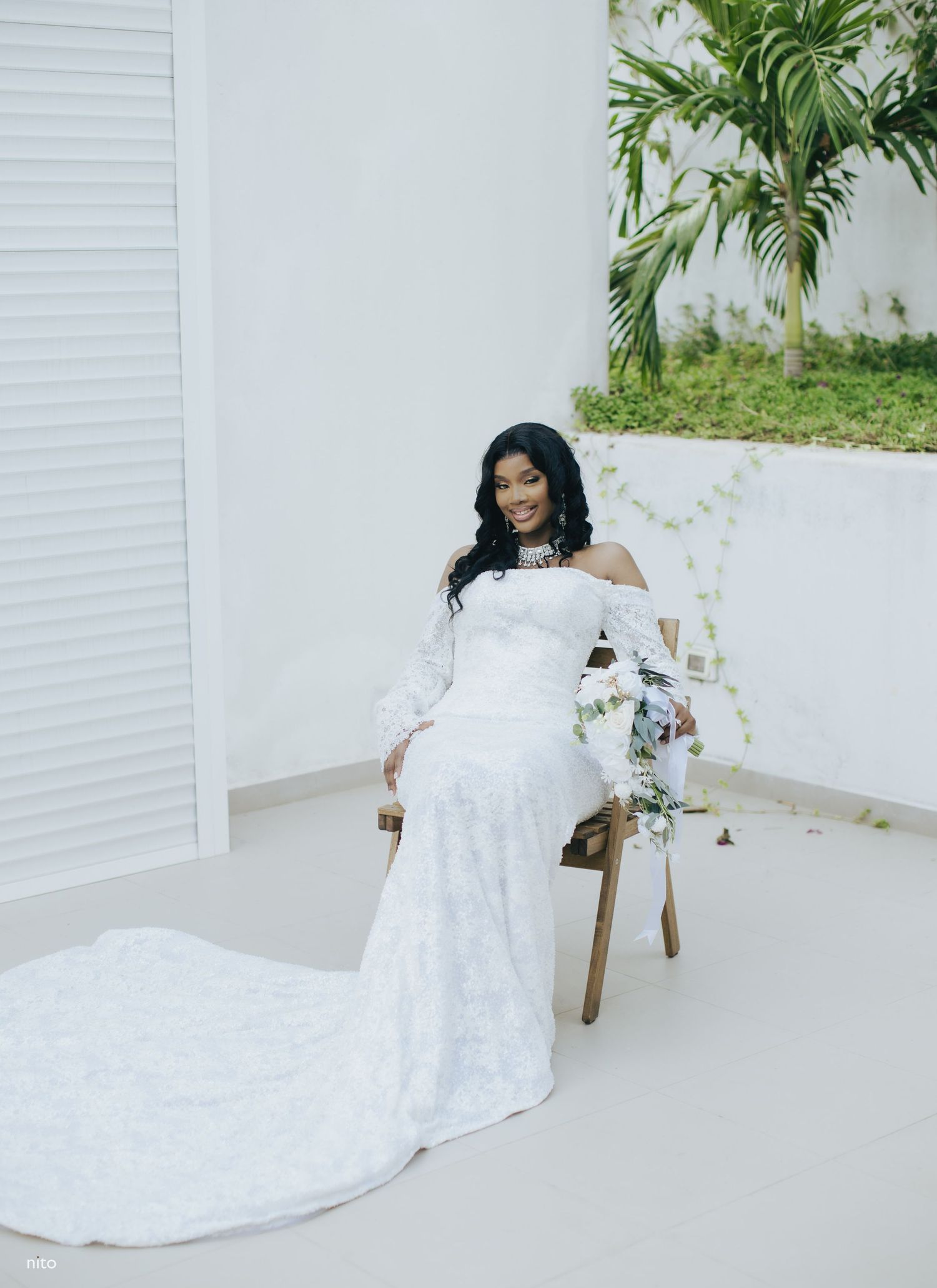A bride in an elegant off-shoulder lace wedding gown with long train sits in a chair against a white wall and tropical palm.
