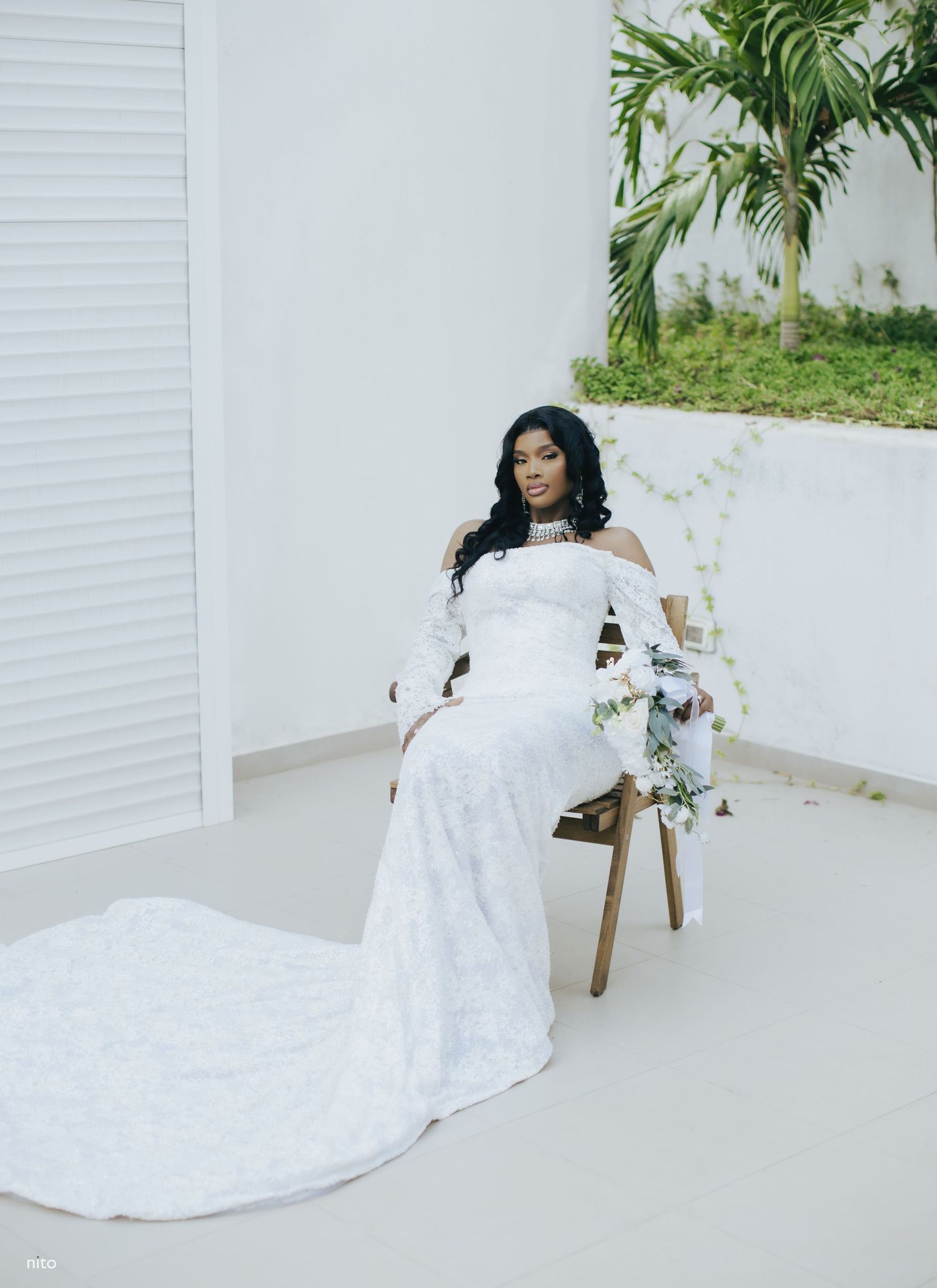A stunning white lace wedding dress with off-shoulder detail and long train displayed in a bright minimal setting with palm trees.
