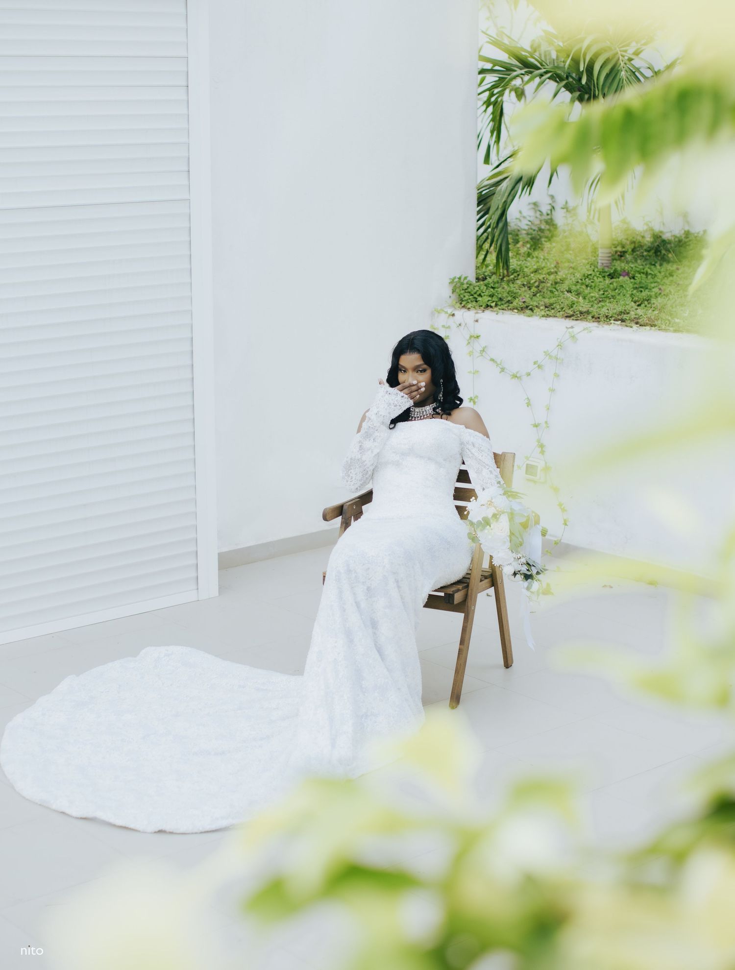 A bride in a flowing white wedding gown with long sleeves sits on a wooden chair against a white wall with tropical decor.
