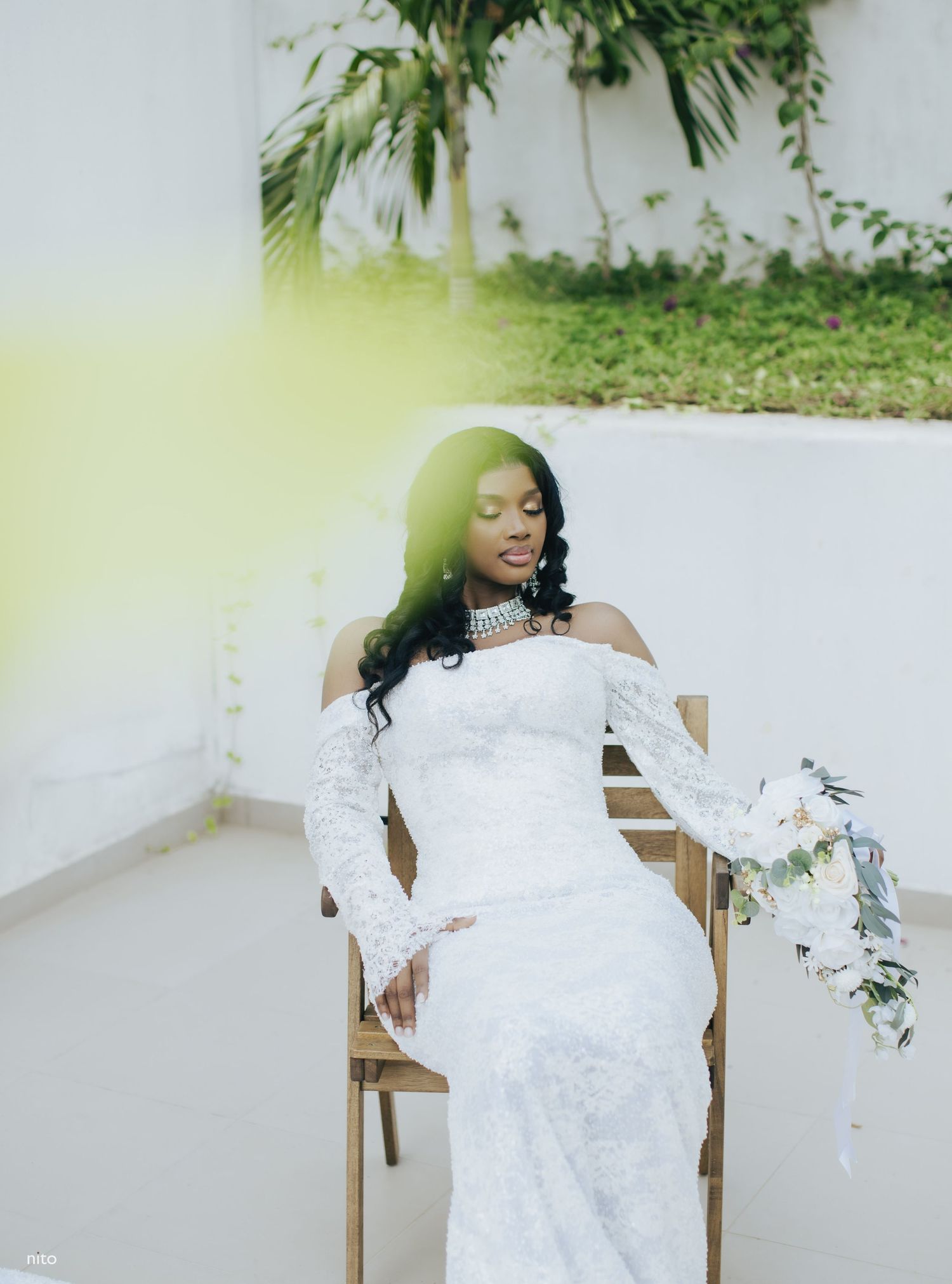 A bride in an elegant off-shoulder lace gown poses gracefully with a white floral bouquet against a light background.