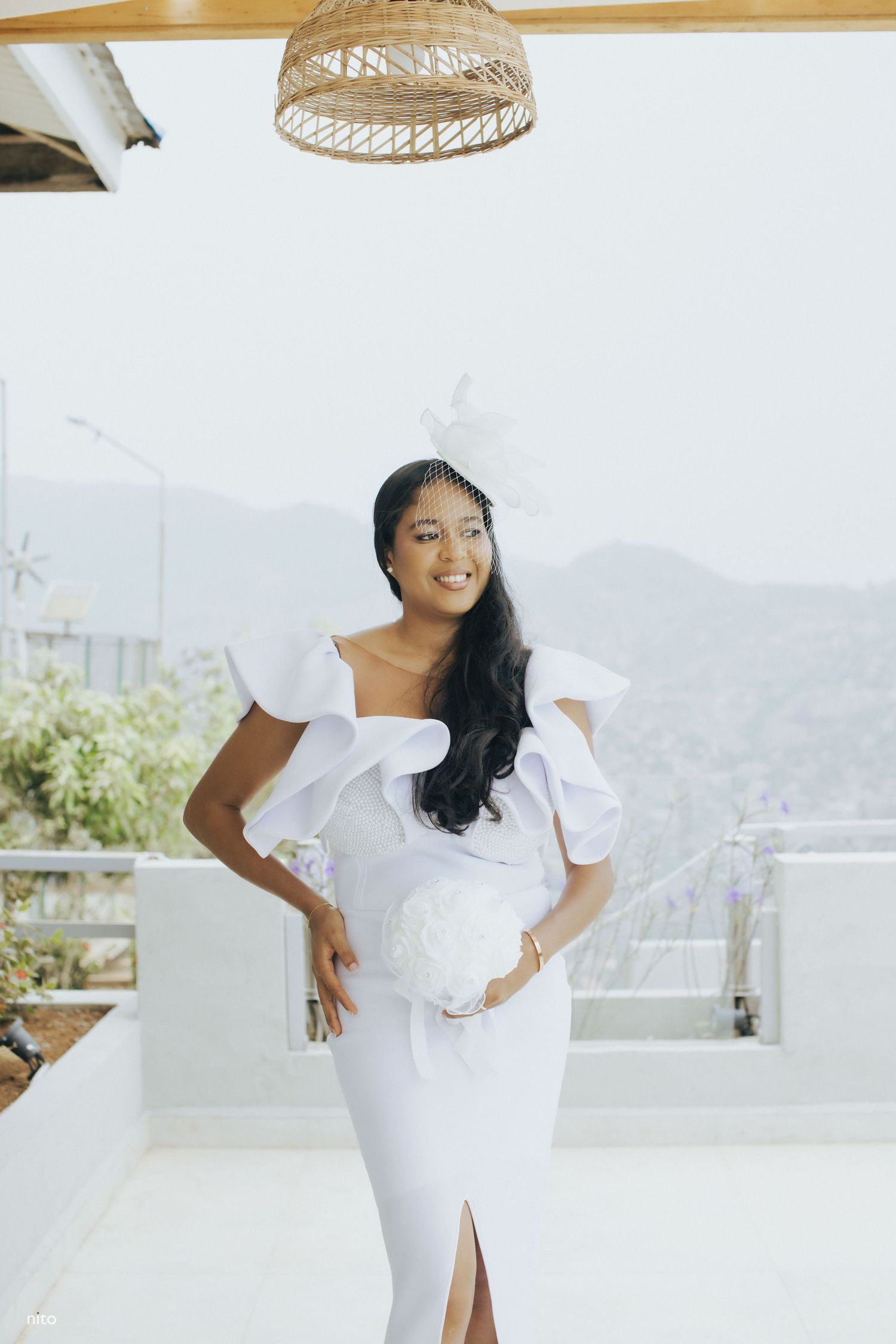 A bride in a sleek white dress poses under a woven pendant light with mountain views.