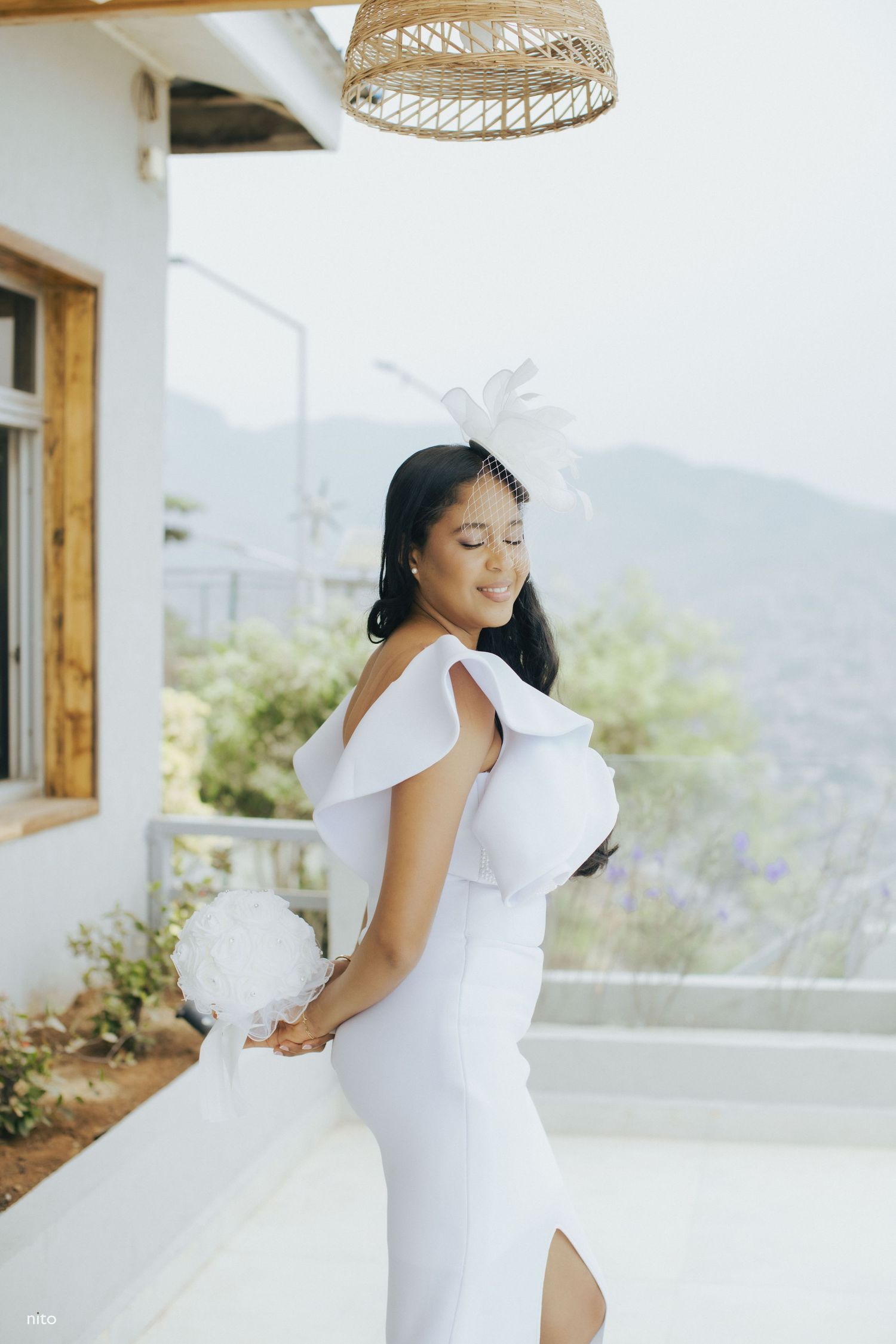 A bride in a modern white dress poses on a balcony with mountain scenery.