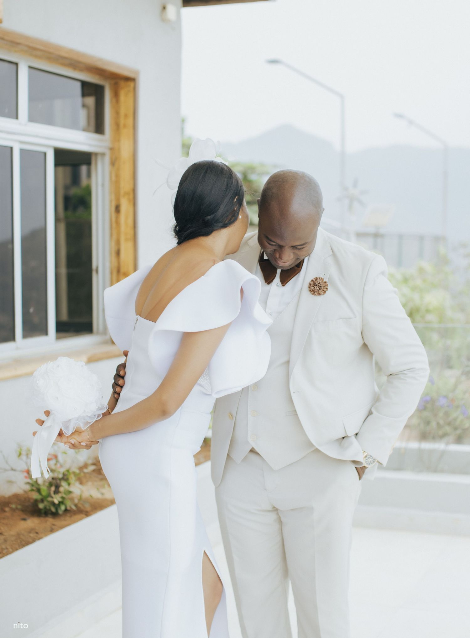 A couple in elegant white attire shares a tender moment on a scenic terrace.