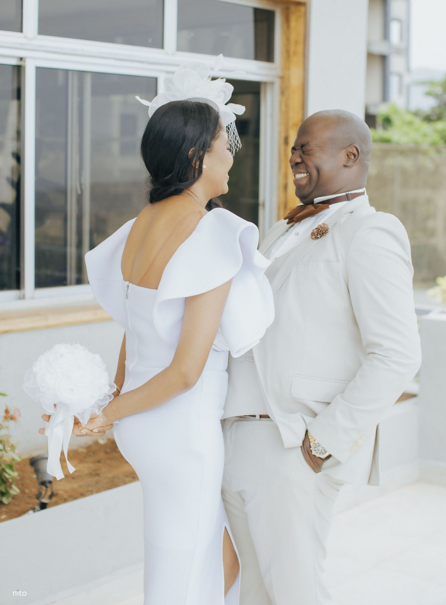 A joyful wedding couple in white attire shares a laugh on their scenic balcony.