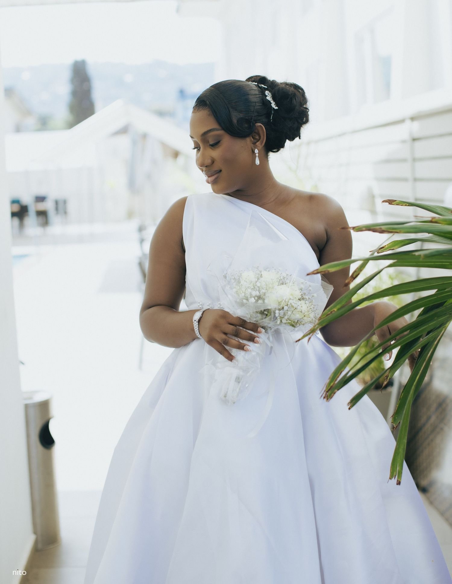 A bride in an elegant one-shoulder white wedding dress holds a delicate bouquet against a light background.
