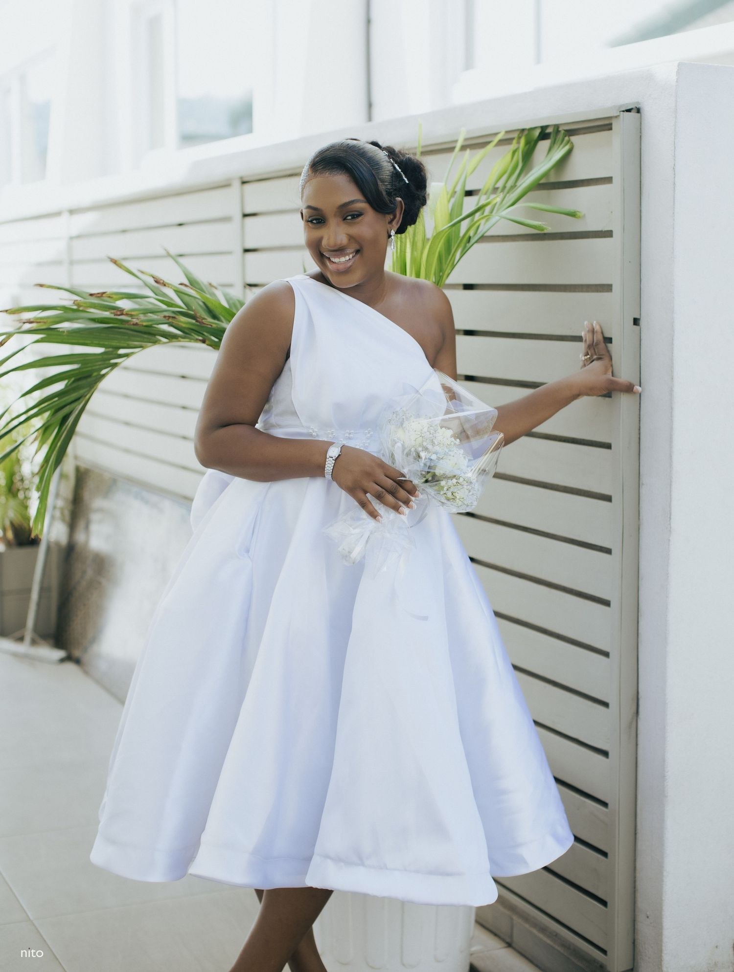 A bride in a tea-length white wedding dress poses with flowers against white shutters.