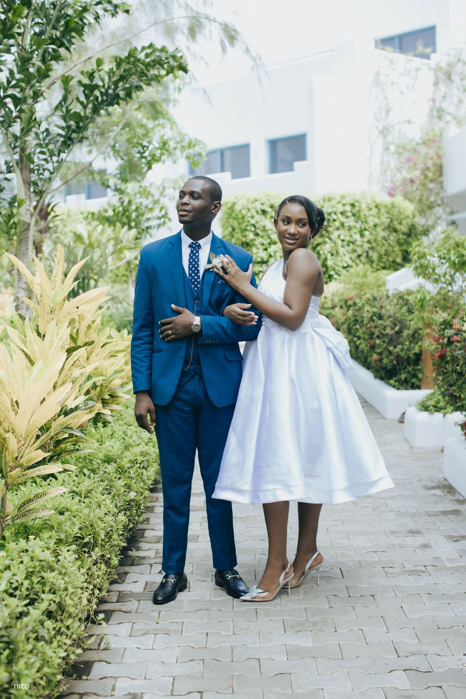 Stylish couple poses on garden pathway in wedding attire.