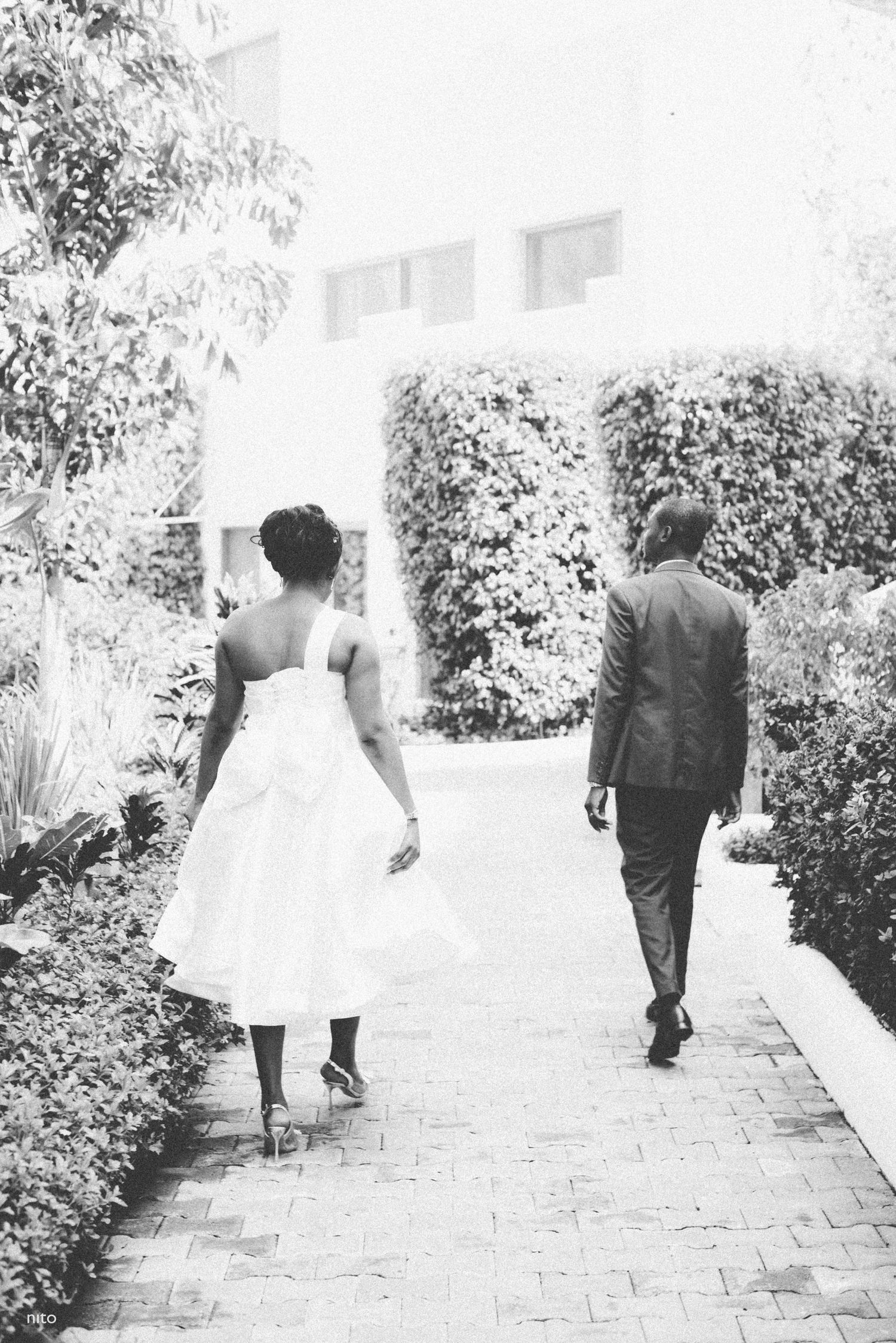 Black and white image of couple walking through garden pathway in wedding attire.