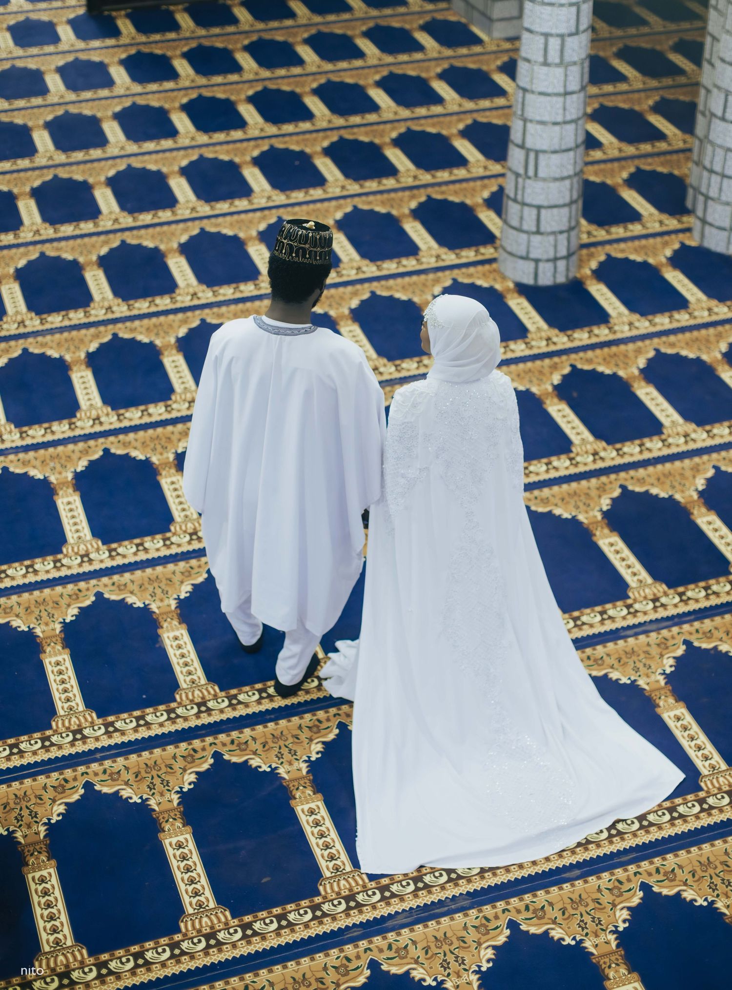 Two people in white garments stand on ornate blue and gold prayer carpets in a mosque.