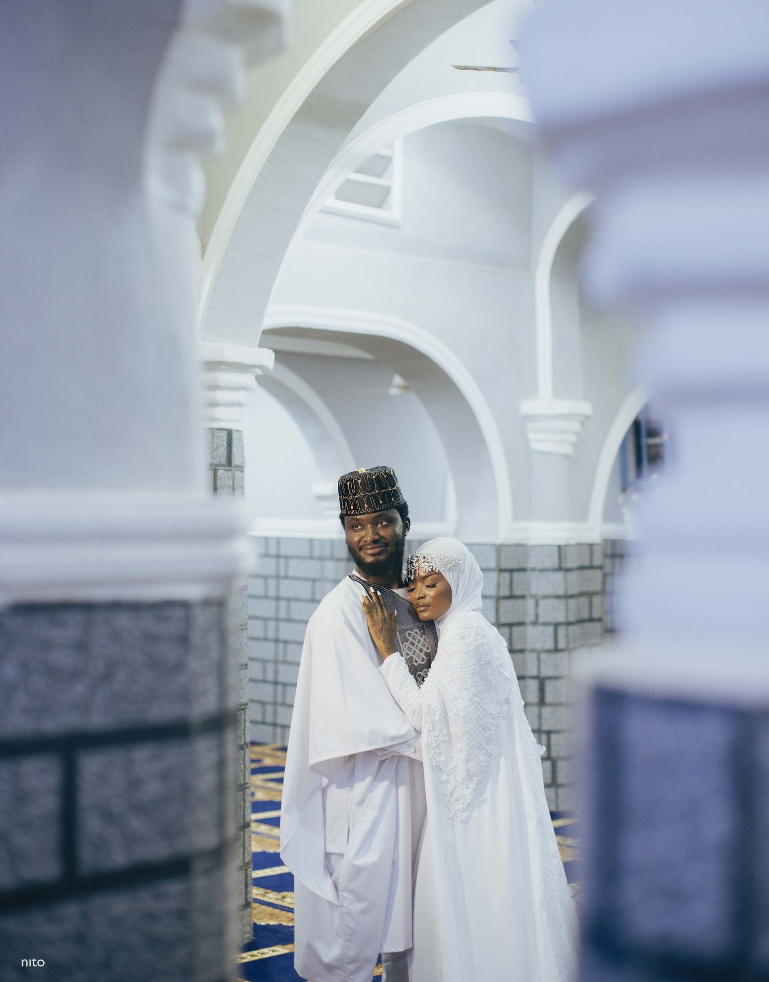 Two people in white religious attire embrace in prayer at a mosque with arched architecture.