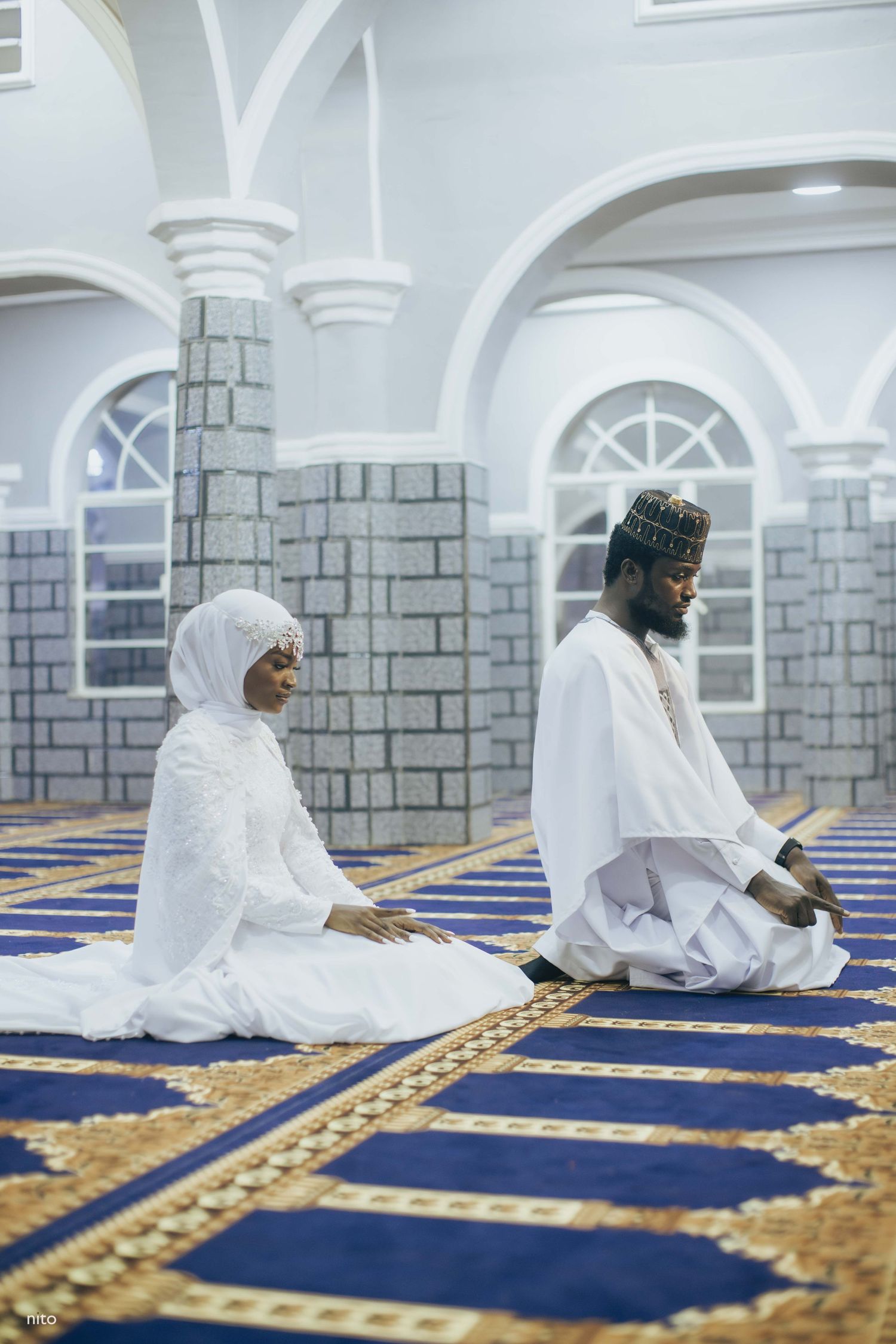 Two people pray on decorated blue and gold carpets inside a mosque with white stone columns.