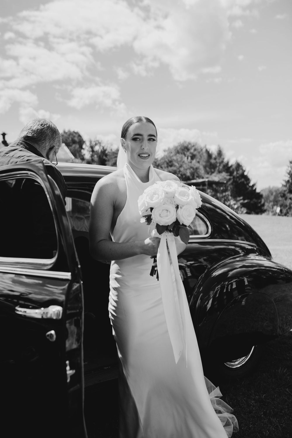 Bride in white halter gown holds white rose bouquet beside vintage black car in black and white.
