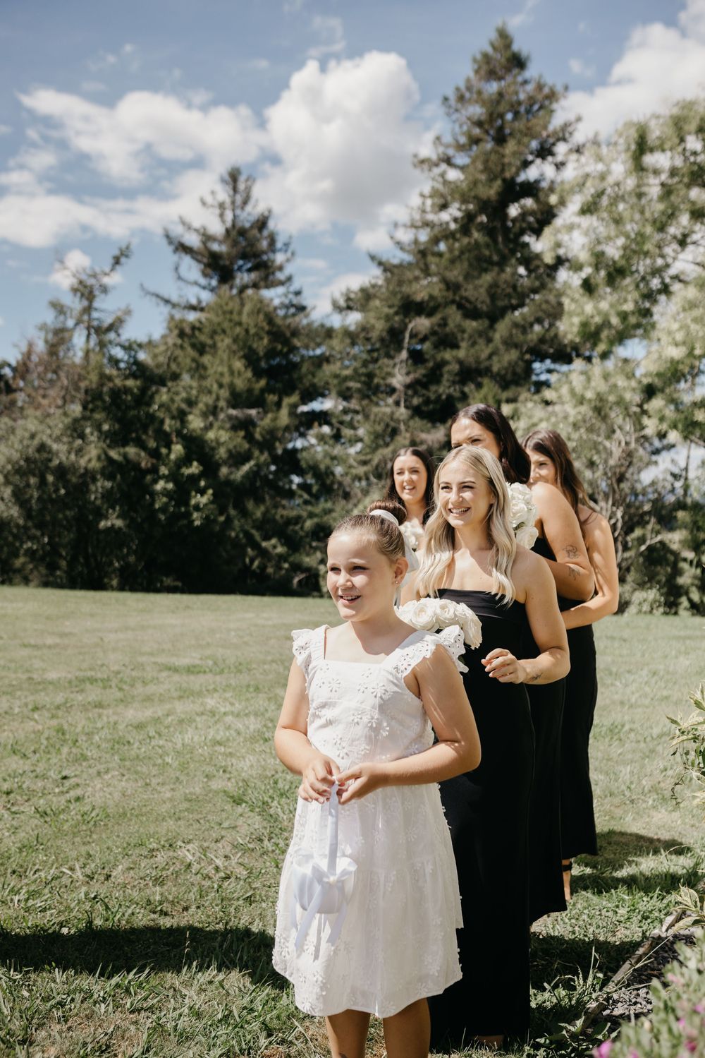 Flower girl in white dress leads bridesmaids in black gowns across green lawn under blue sky.