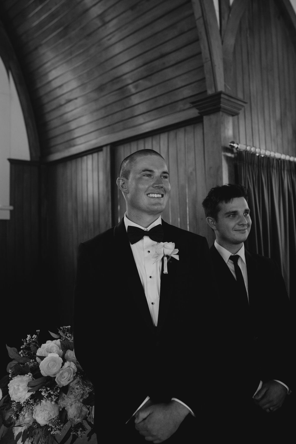Smiling groom in black tuxedo stands at altar with groomsman in black and white church photo.