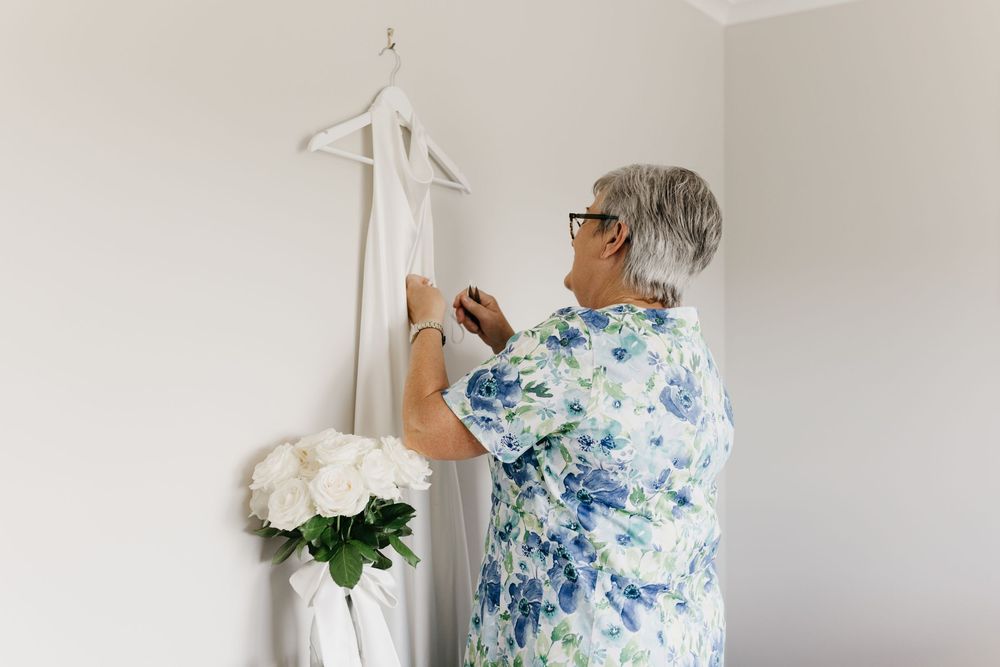 Mother of the bride carefully arranges elegant white wedding gown on hanger with white rose bouquet nearby.