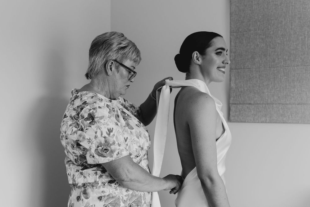 Black and white photo of mother helping bride fasten her elegant halter neck wedding dress.