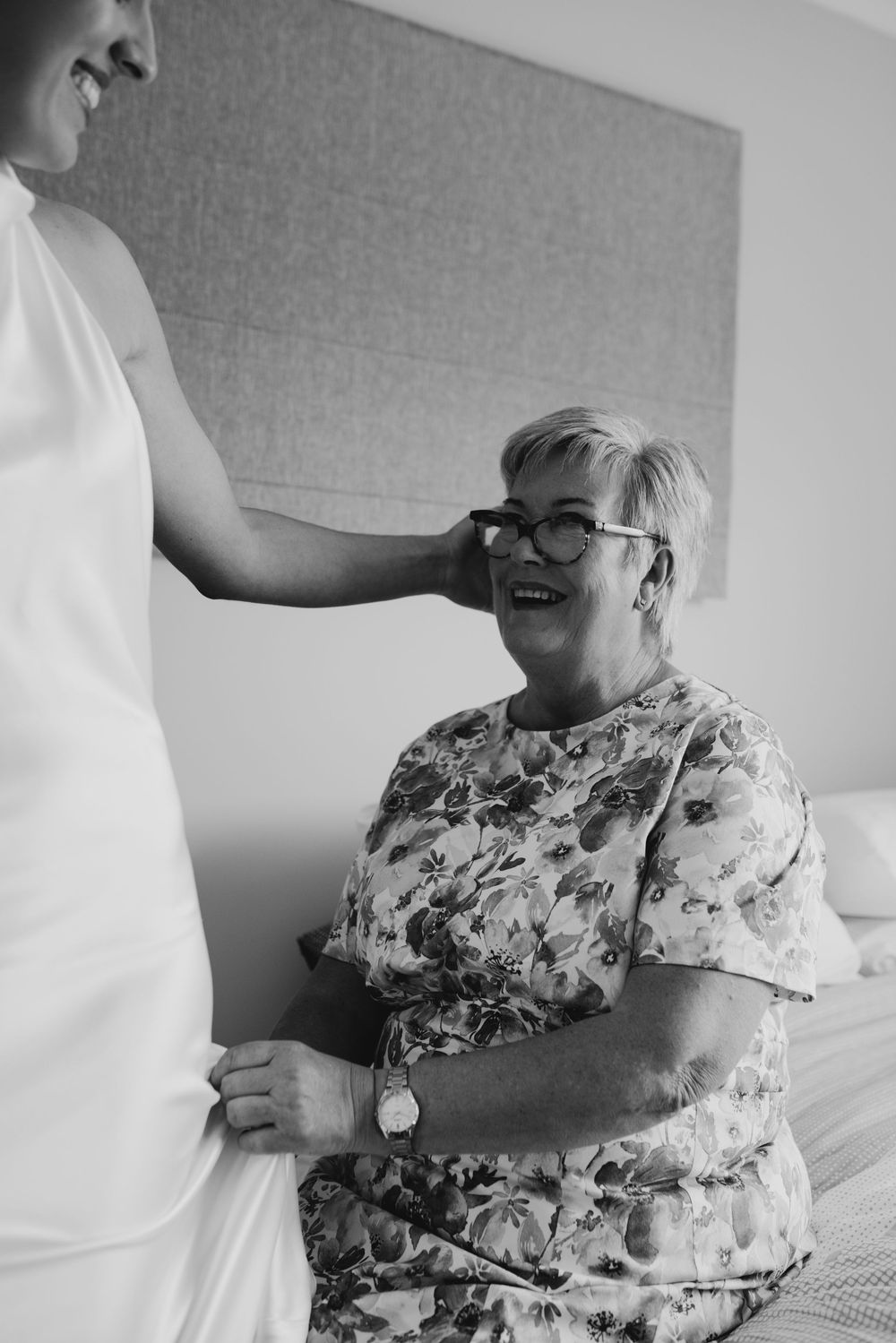 Black and white photo of smiling mother sitting on bed watching bride get ready for wedding day.