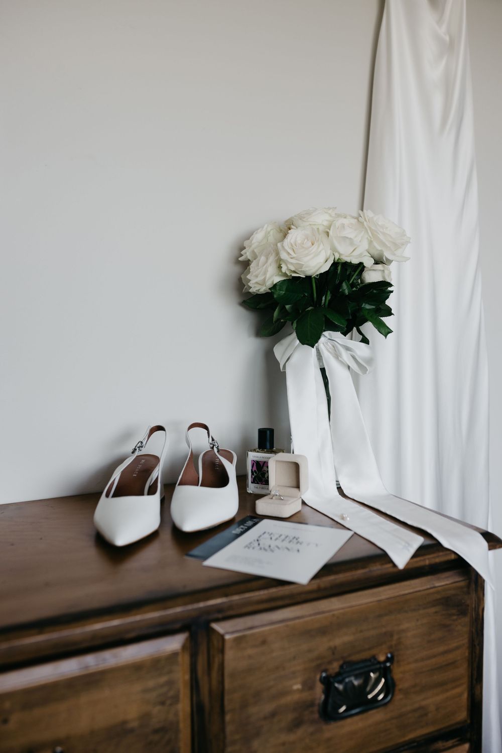 Bridal flat shoes, white rose bouquet, perfume, and ring box arranged on a rustic wooden dresser.
