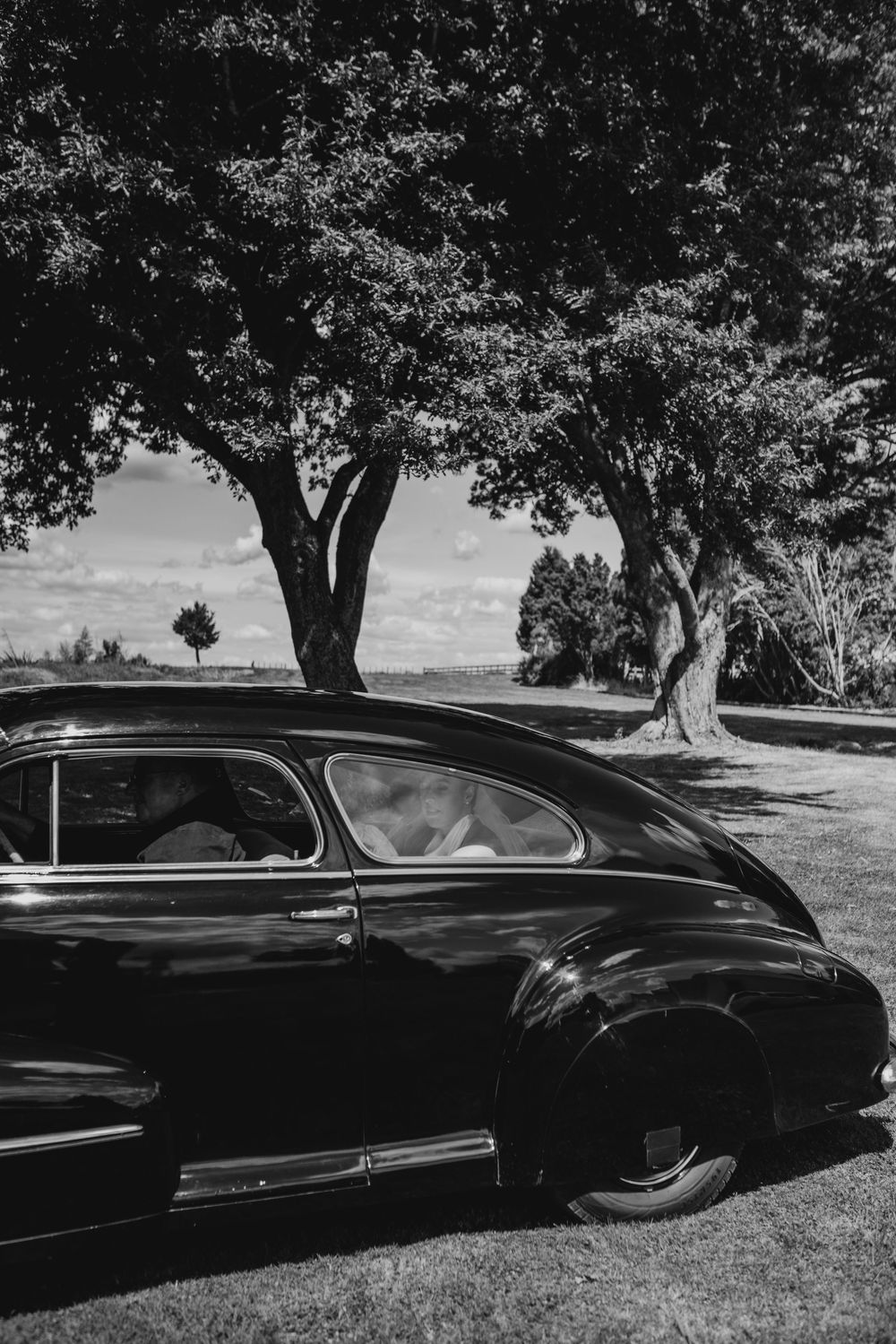 Vintage black classic car parked under large shade trees at an outdoor wedding venue.