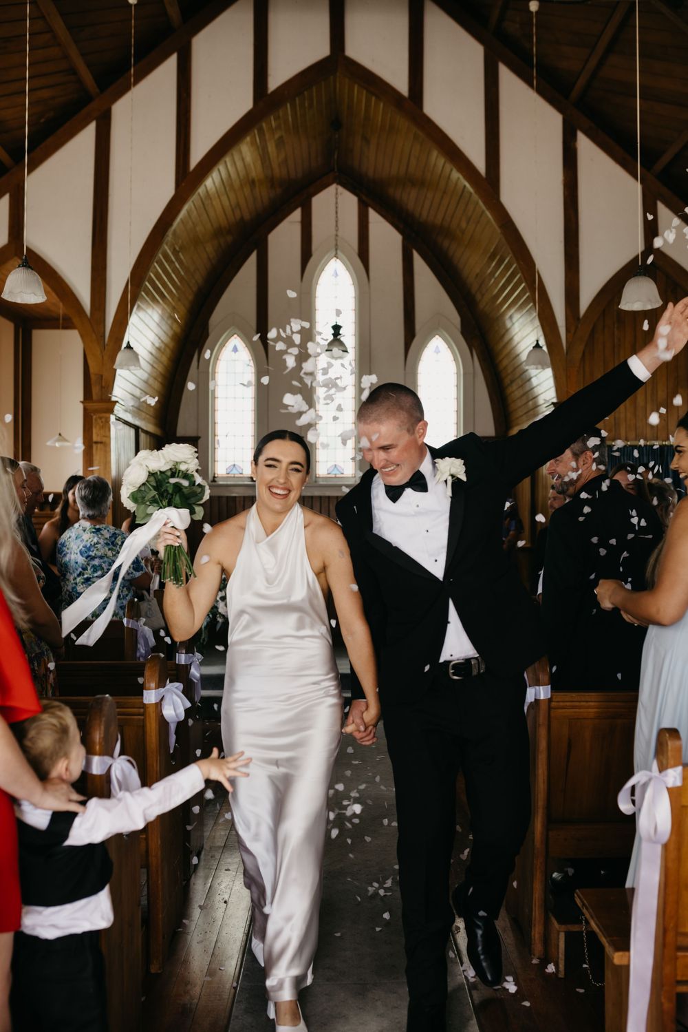 Joyful newlyweds walk down church aisle as guests toss white confetti during wedding recessional.