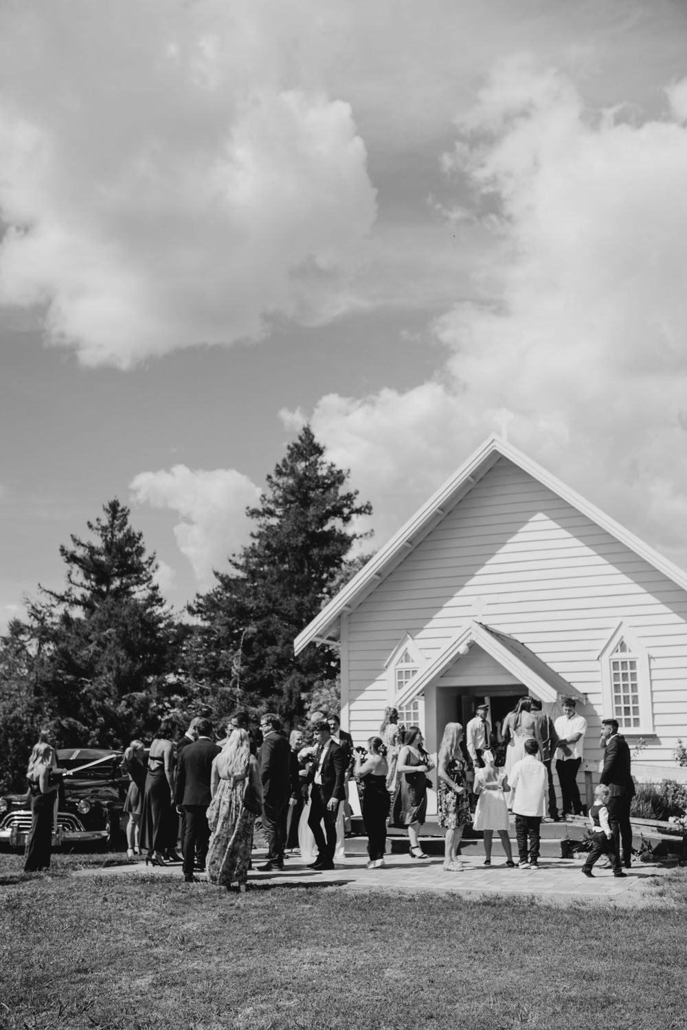 Black and white photo of wedding guests gathering outside a white country church on a sunny day.