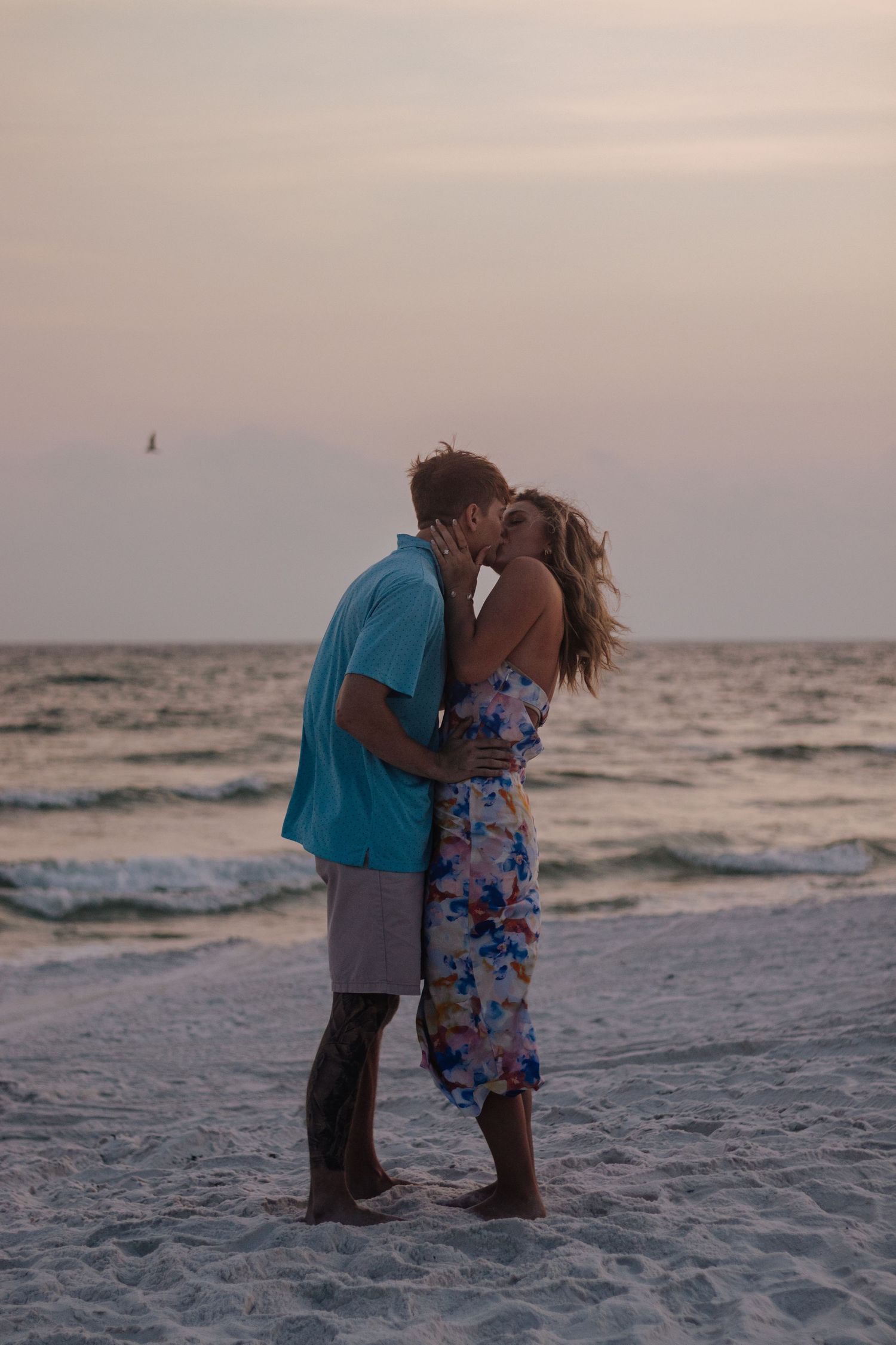 A romantic couple shares a tender kiss on a beach at sunset with waves crashing in the background.