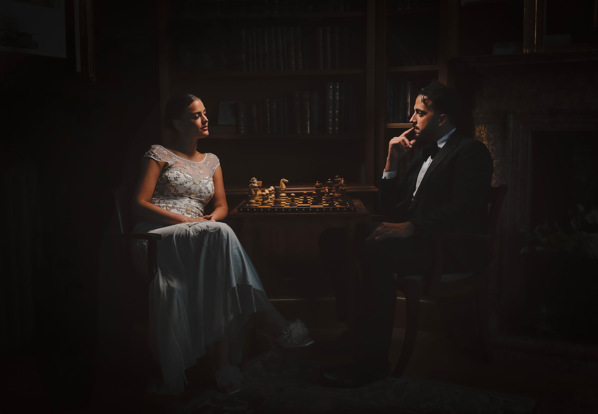 Bride and groom playing chess by candlelight in a dark, elegant library setting.