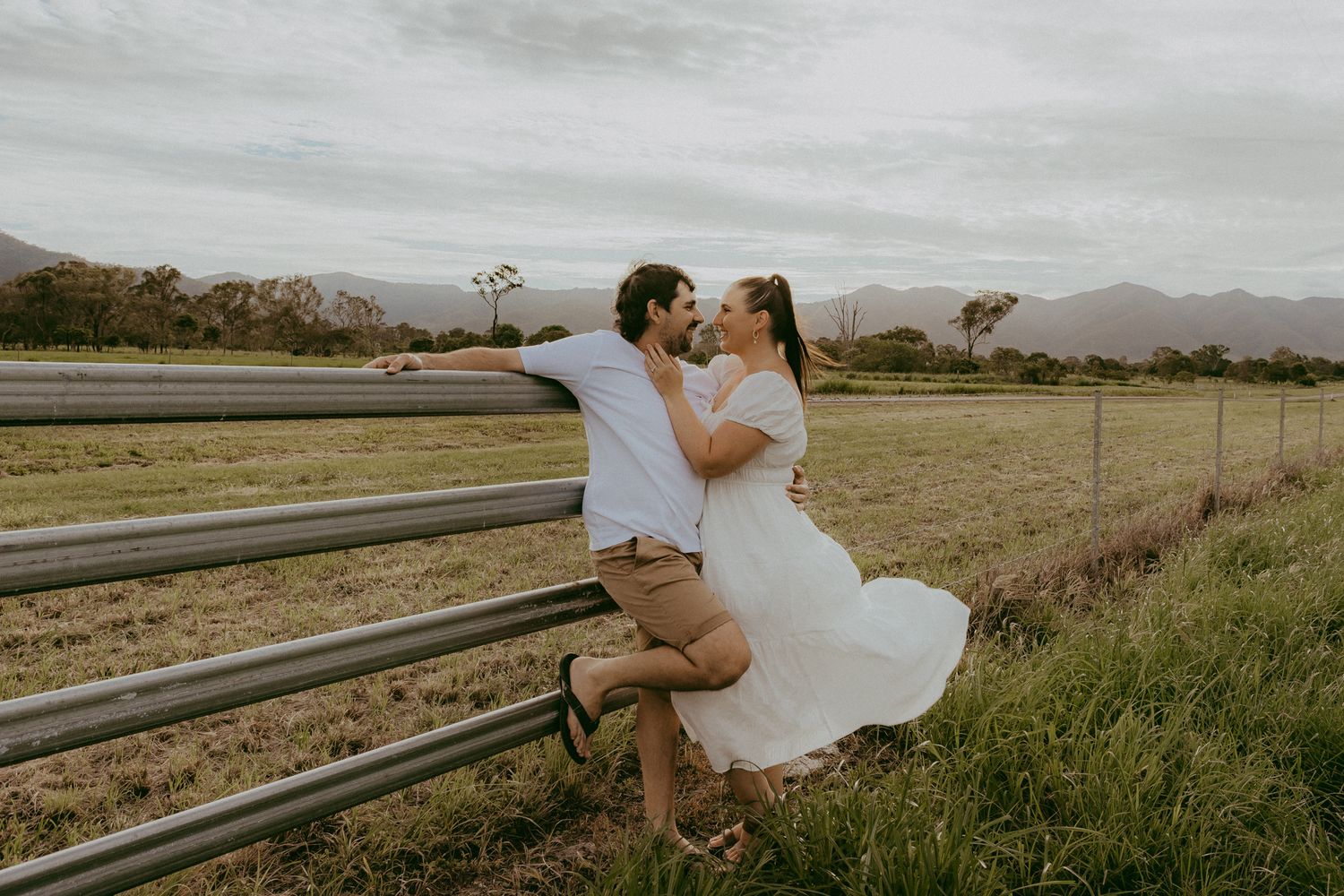 Couple sharing a romantic kiss leaning against a metal railing in a scenic countryside setting with mountains in the background.